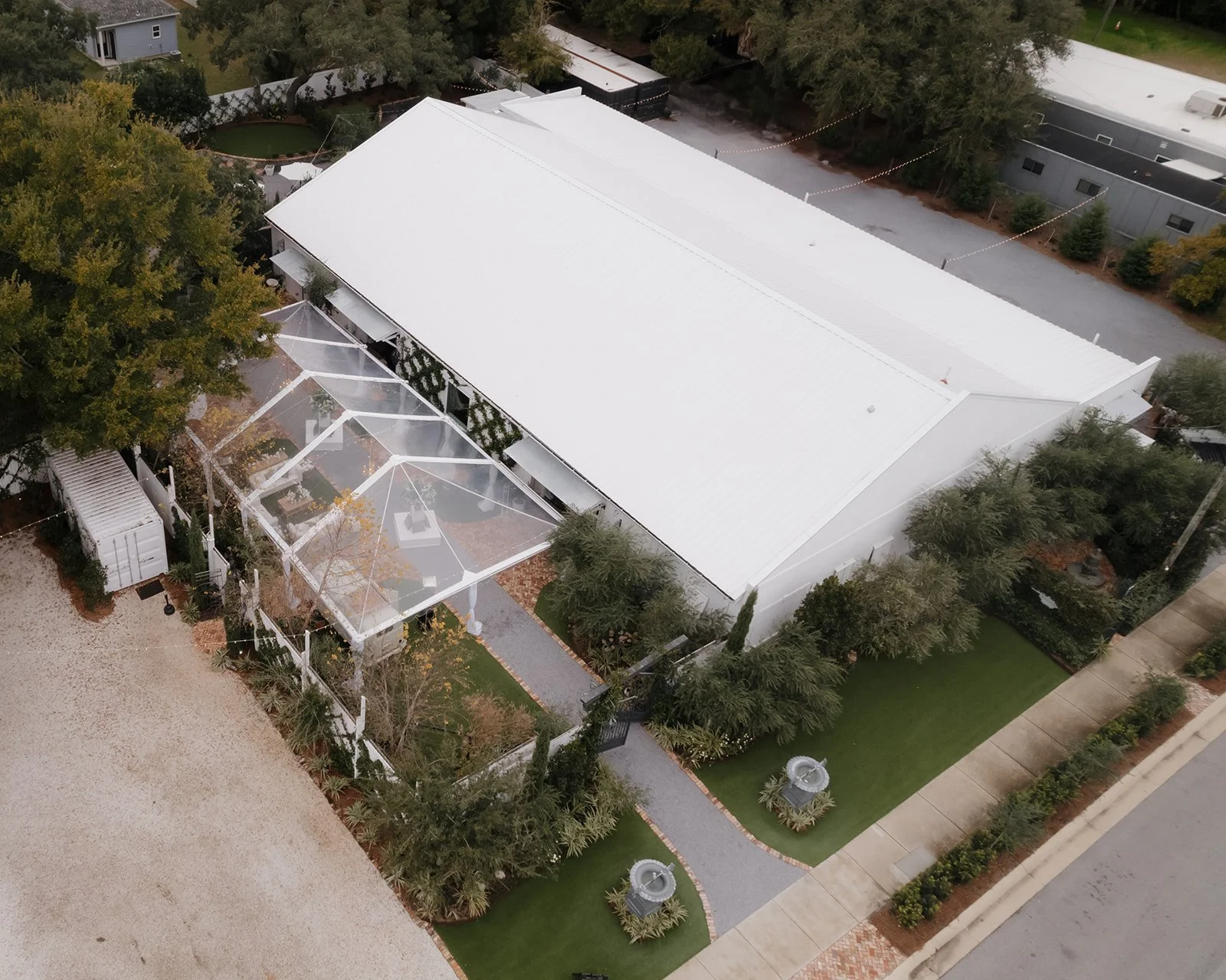 Aerial view of a large white wedding venue with a metal roof, adjacent to a landscaped yard with trees, a gravel area, and a walkway. A clear tent enclosed patio is attached to the building, and there are decorative fountains near the sidewalk.