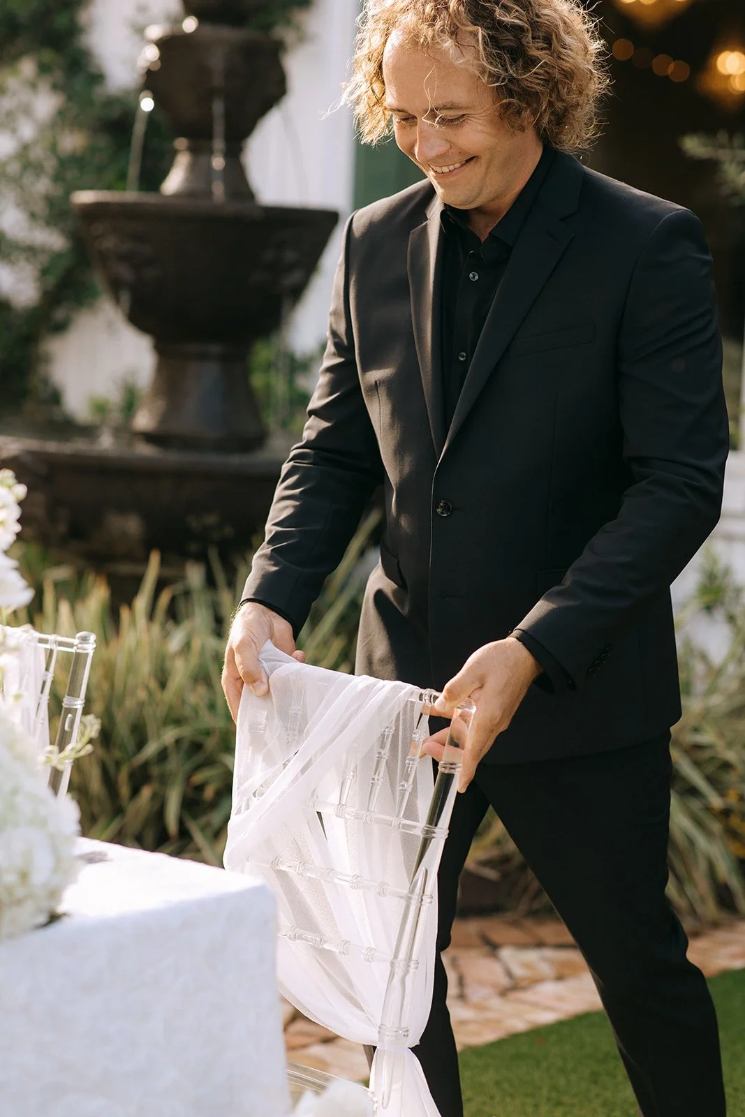 A Wedding Venue Owner in a black suit adjusting a white chair at an outdoor event, smiling as he prepares a table with a wedding cake in the foreground.