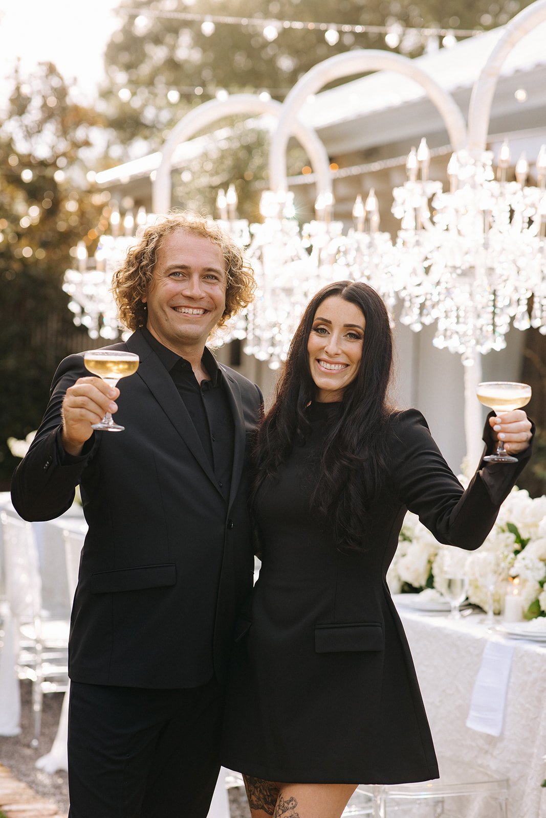 Pensacola Wedding Venue Owners dressed in black, smiling and holding glasses of champagne at an outdoor celebration with chandeliers and white flower decorations.