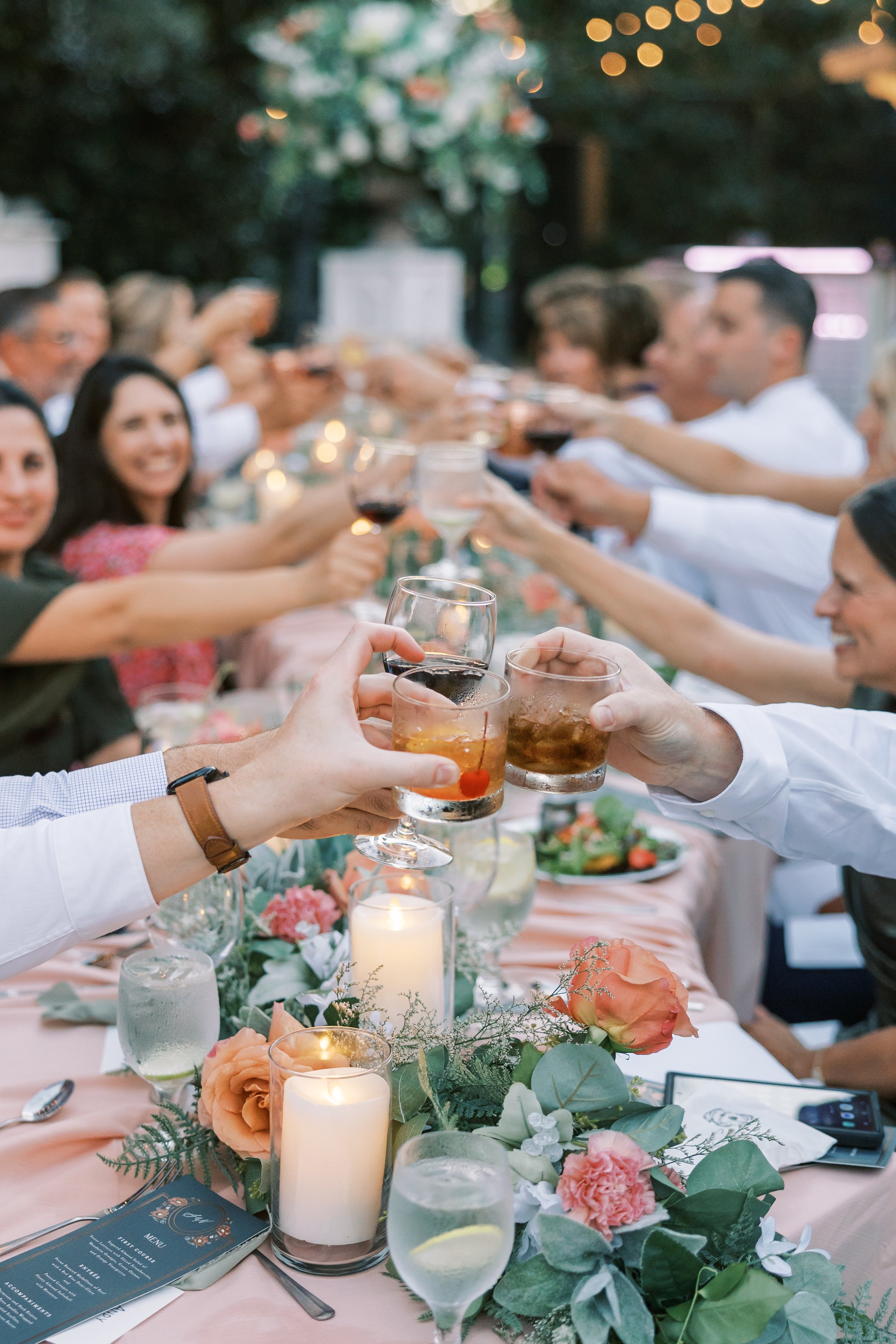 A group of people raising glasses in a toast at a dinner party outdoors with a long table decorated with flowers and candles in a Pensacola Wedding Venue.