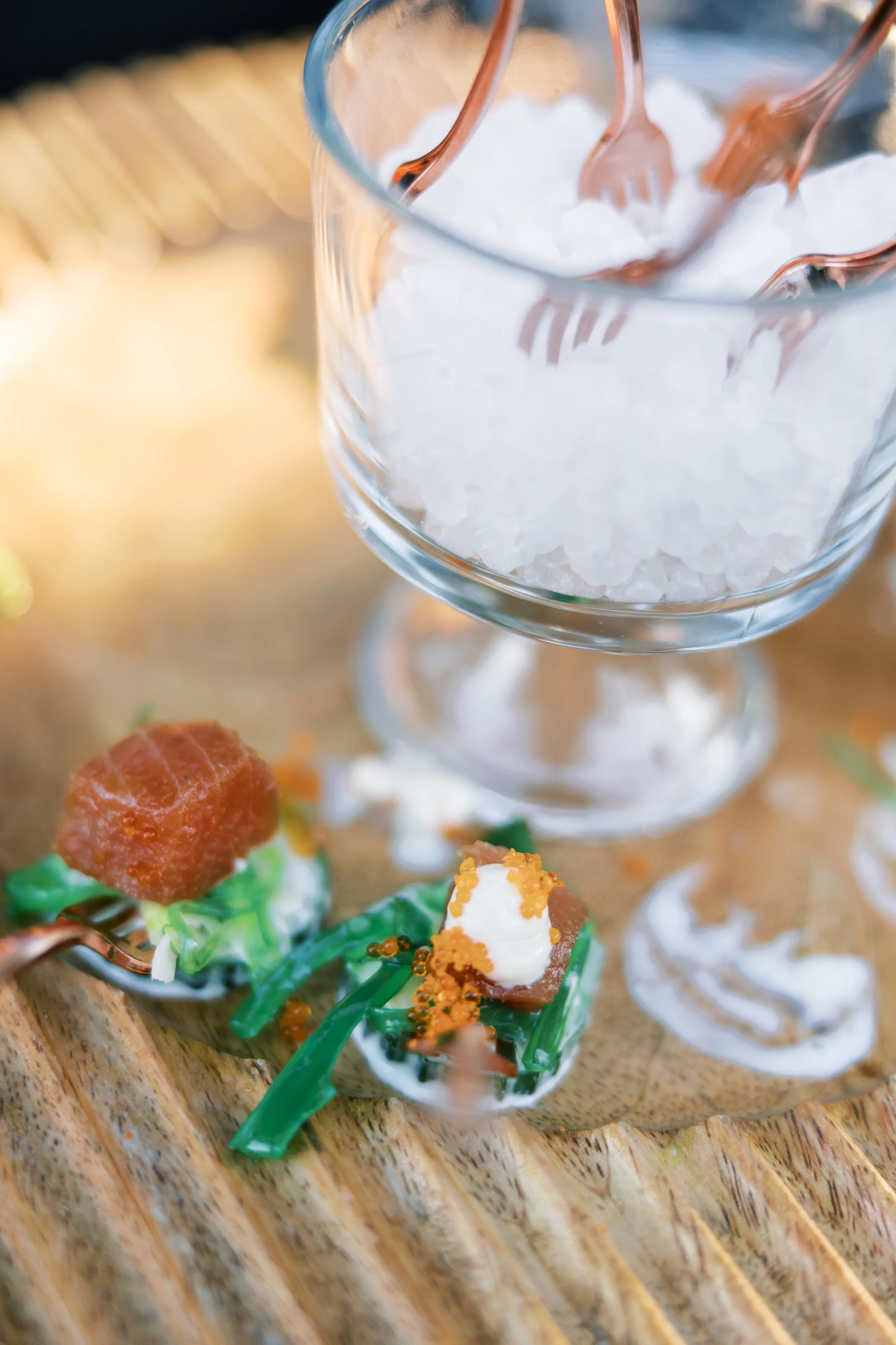 Close-up of a glass bowl filled with ice and three small forks inserted into the ice, with some appetizers on a wooden surface below.