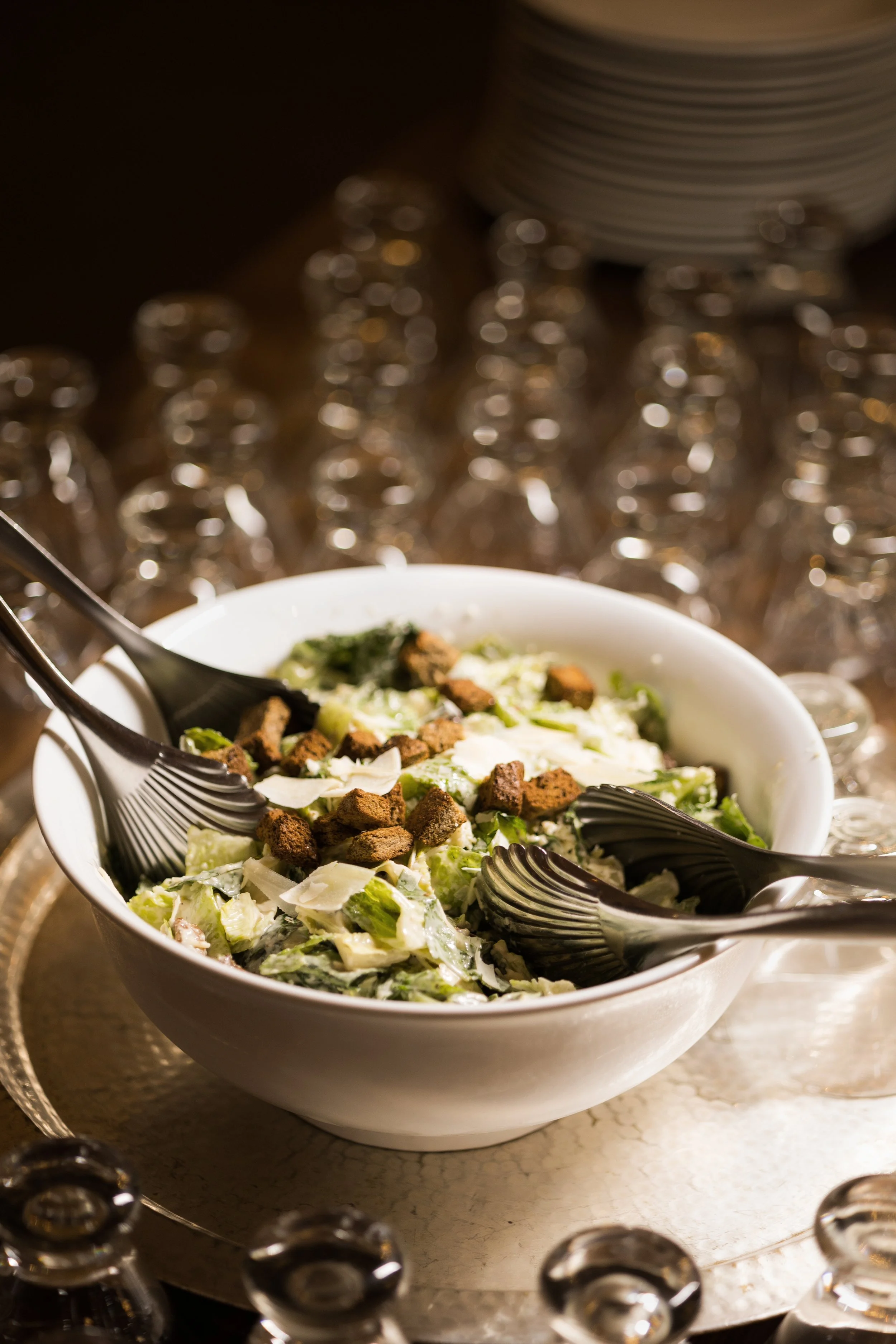 Caesar salad in a white bowl with romaine lettuce, croutons, and shaved cheese, with salad servers inside.