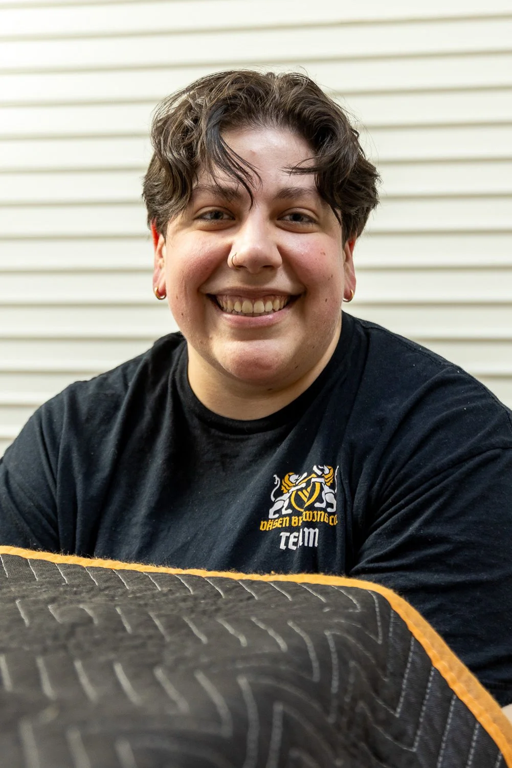 A smiling person with short, wavy dark hair, wearing earrings and a nose ring, dressed in a black T-shirt with a logo, sitting in front of a horizontal beige siding background.