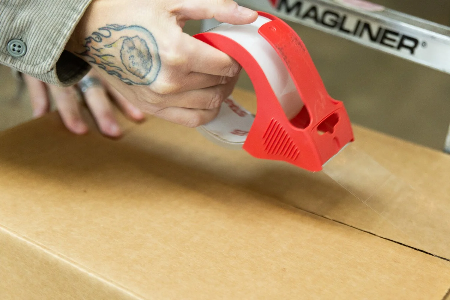 A person using a red tape dispenser to cut a strip of clear packing tape onto a cardboard box.