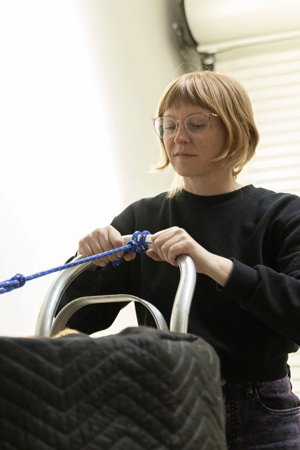 A woman with short red hair and glasses, wearing a black sweatshirt, is tying a knot in a blue rope attached to a cart with a black quilted cover.