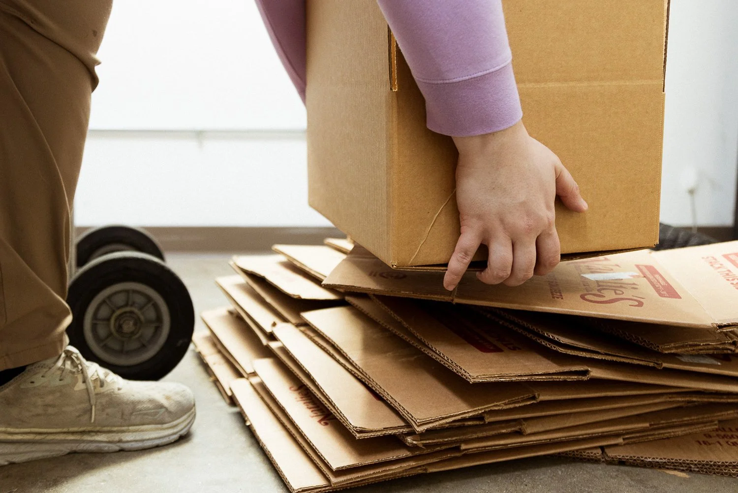 A person wearing beige pants and white sneakers stacking a cardboard box on top of flattened cardboard boxes on the floor, with wheels visible on a dolly in the background.