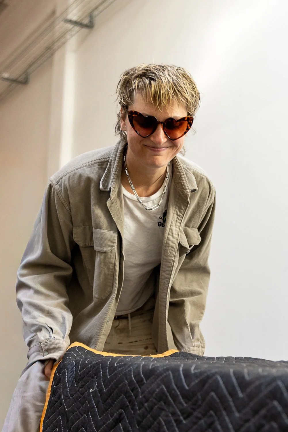 A person with short, blonde hair wearing sunglasses, a beige jacket over a white T-shirt, and a silver necklace, leaning forward and smiling.