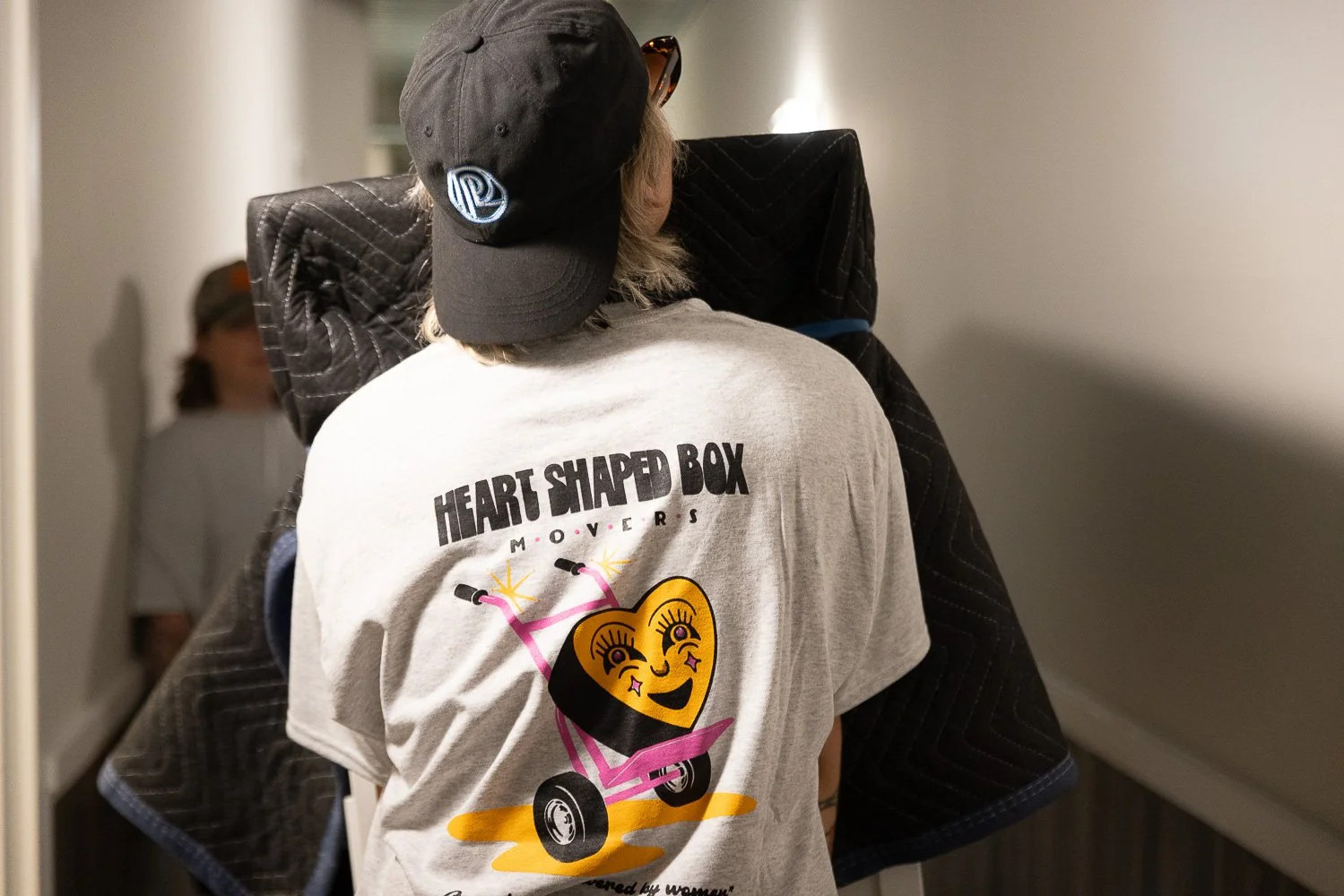 Back view of a person wearing a gray T-shirt with a heart-shaped character on a skateboard and the text 'Heart Shaped Box Movers'. The person is also wearing a black cap and sunglasses, with another person partially visible in the background.