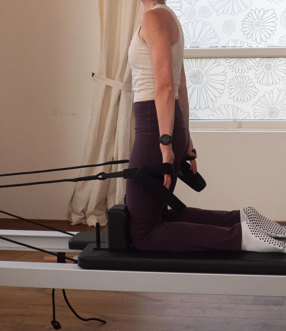 Persoon ligt op een pilates- of fitnessapparaat met weerstandbanden, in een kamer met raam en bloemenpatroon. Het is een man of vrouw, zittend op knieën.