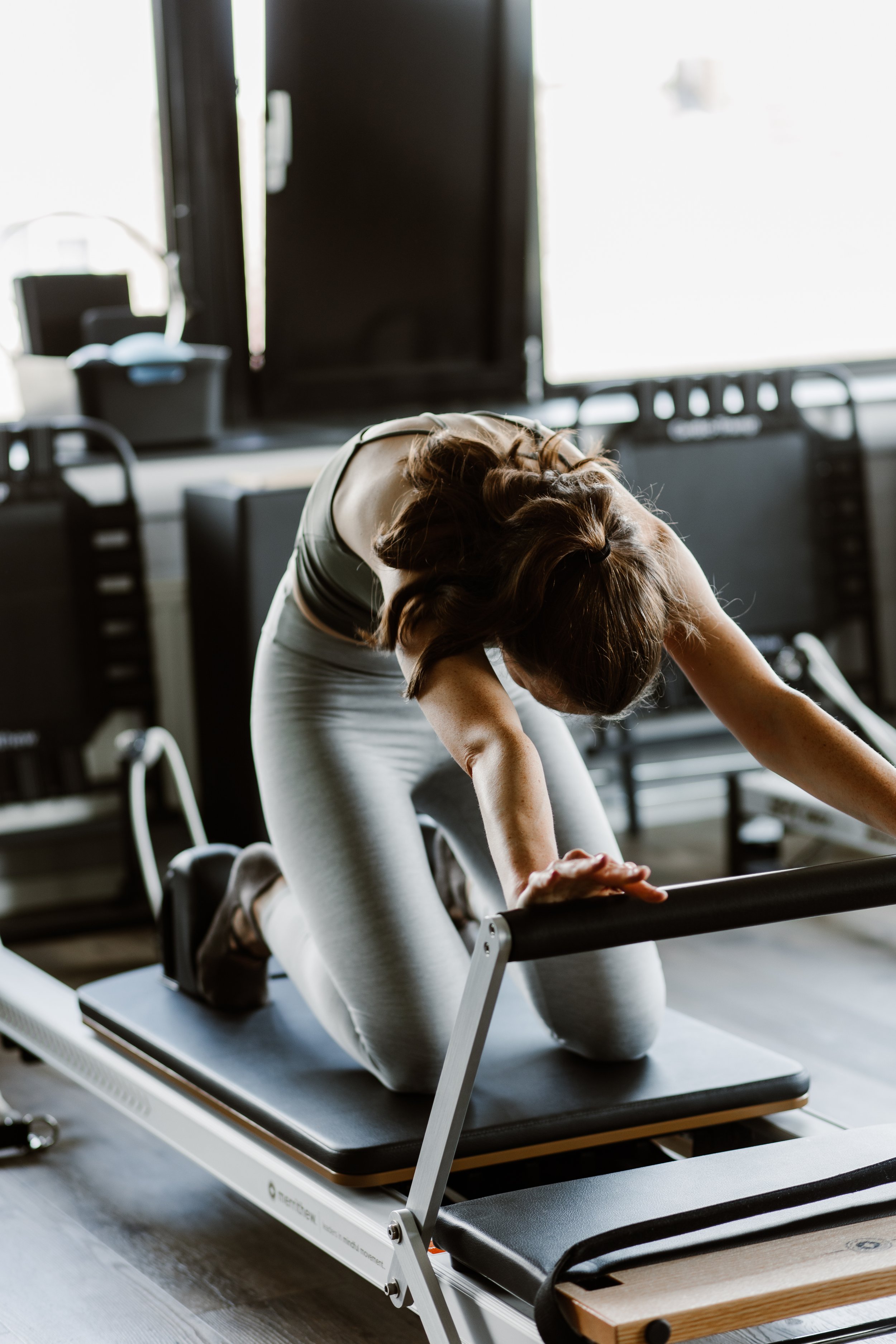 Vrouw die op haar knieën op een fitnessbank aan het stretchen is in een sportschool met veel natuurlijk licht.