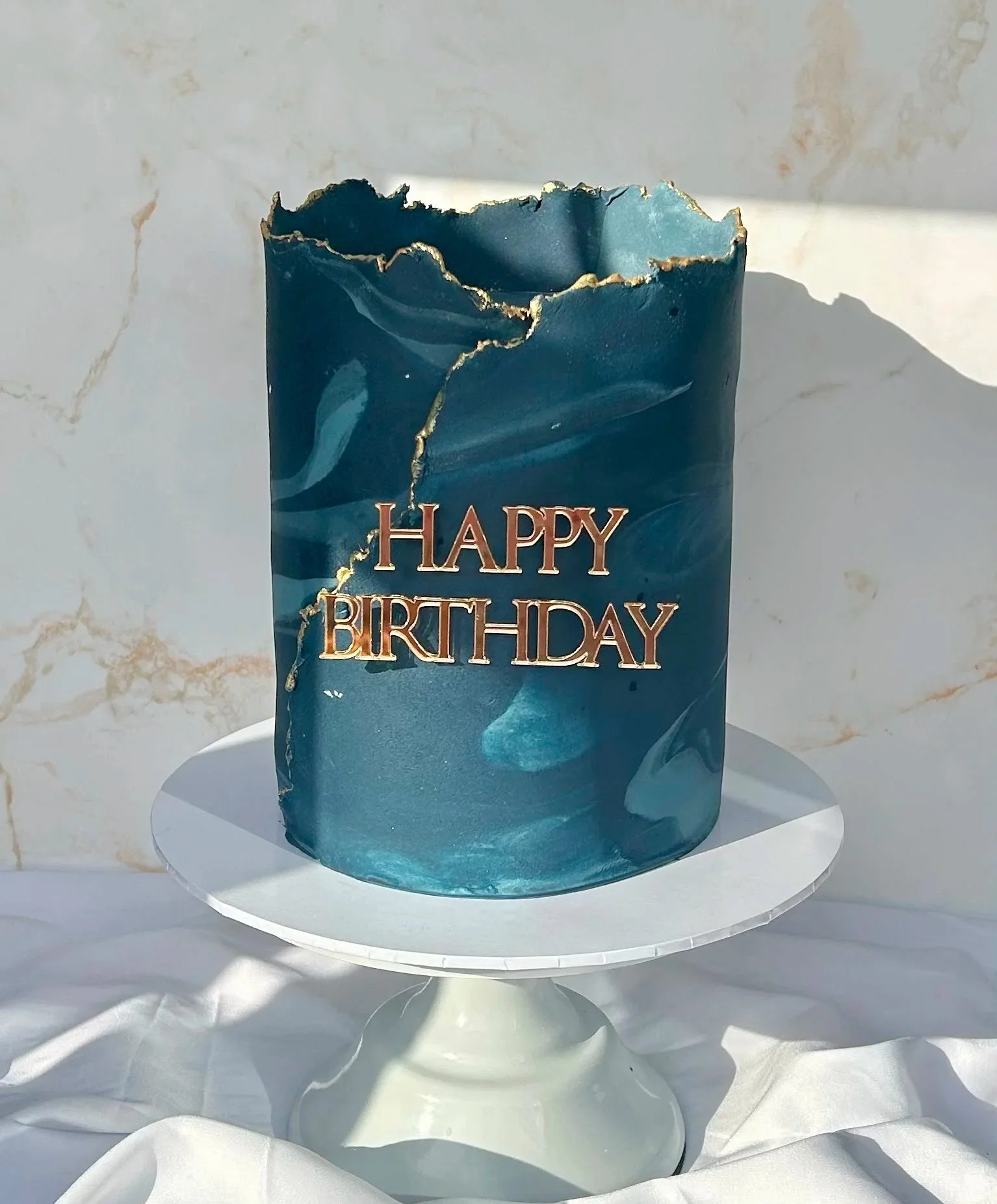 A blue birthday candle with a gold-trimmed, irregular top, placed on a white circular stand. The candle is against a white marble background with subtle gold veining.