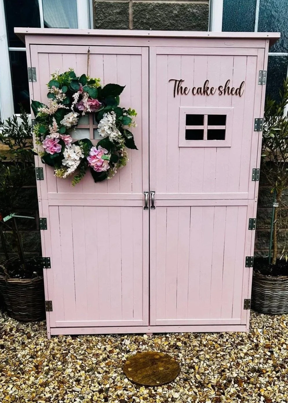 A pink wooden storage shed labeled "The cake shed" with a decorative floral wreath hanging on the left door. The shed has a small window on the right door, metal hinges, and is set on a gravel surface.