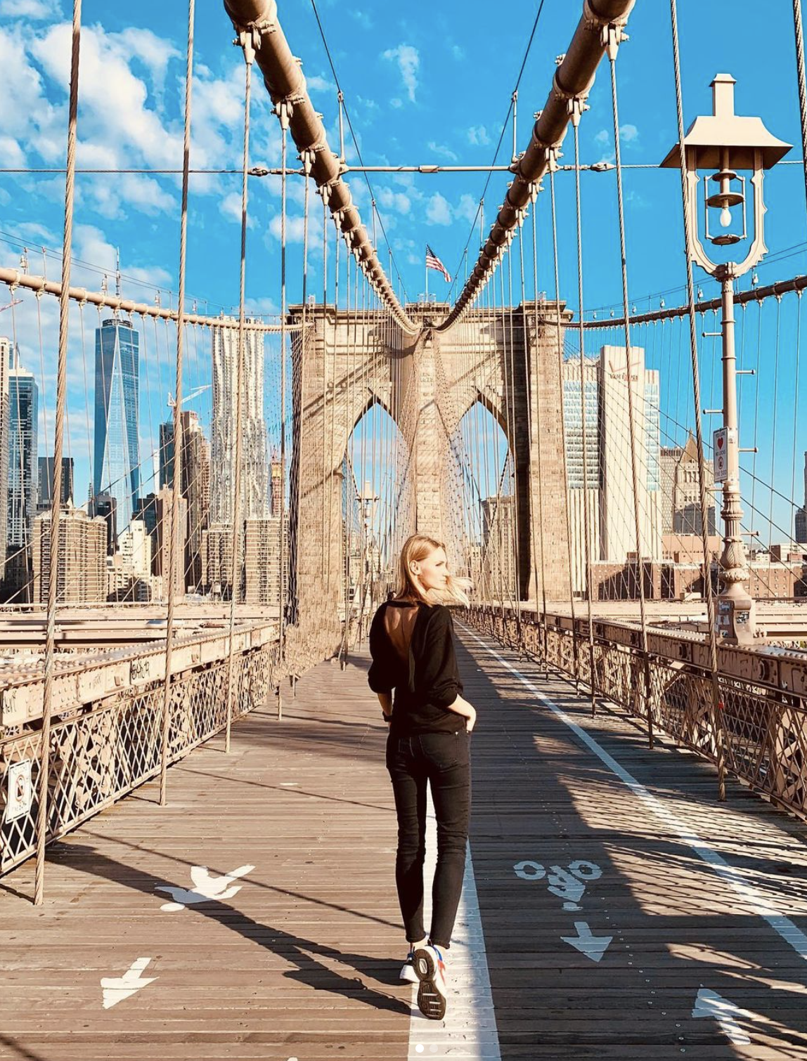 A fitness trainer walking on the Brooklyn Bridge with Manhattan skyline in the background, under a blue sky with some clouds.