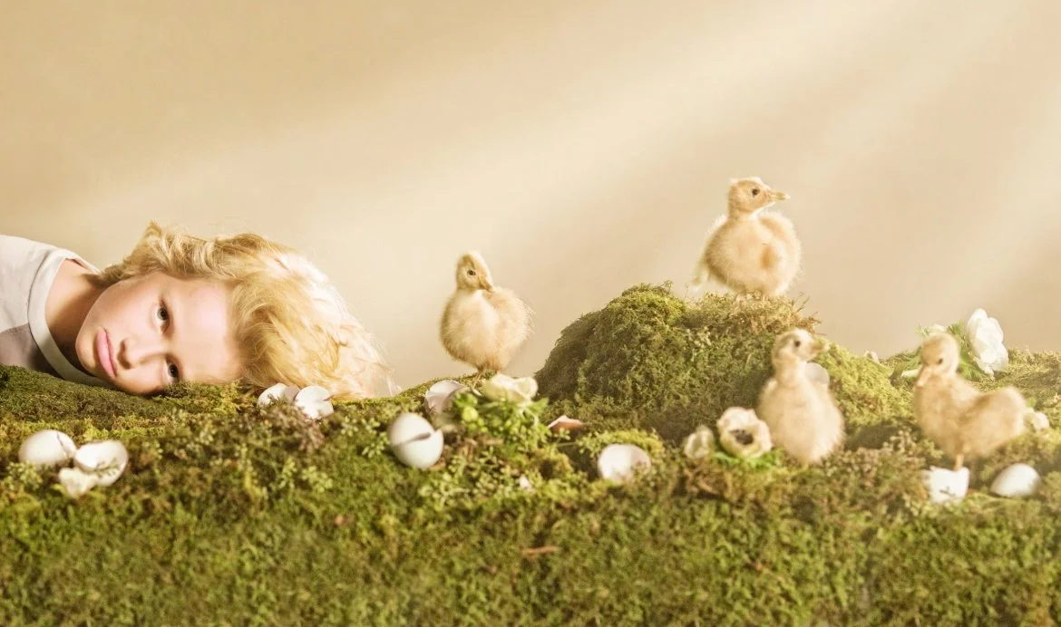 A person with a light-colored wig resting their head on a mossy surface next to young ducklings and cracked eggshells in a natural setting.