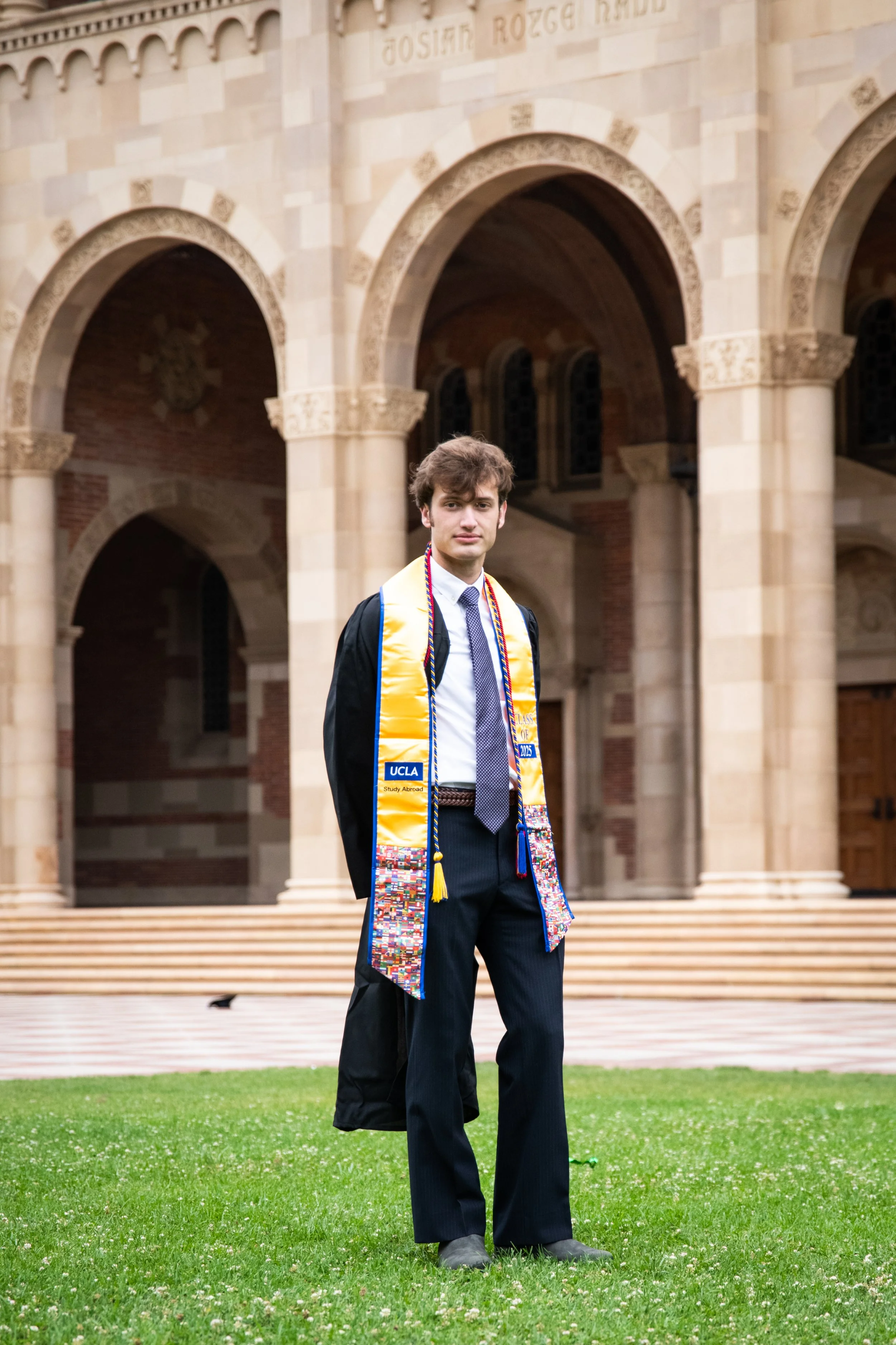 A young man in a black graduation gown and cap, standing on a grass lawn in front of a large, historic building with arched doorways. He is wearing a colorful graduation stole and cords, and looking at the camera.