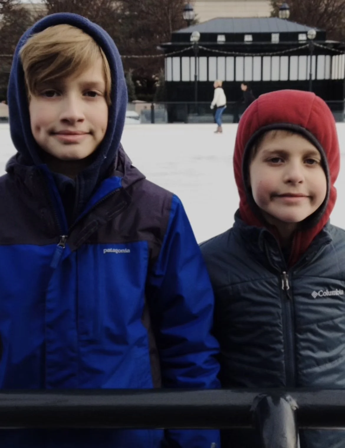 Two boys wearing hooded jackets standing outdoors near an ice skating rink with a pavilion in the background.