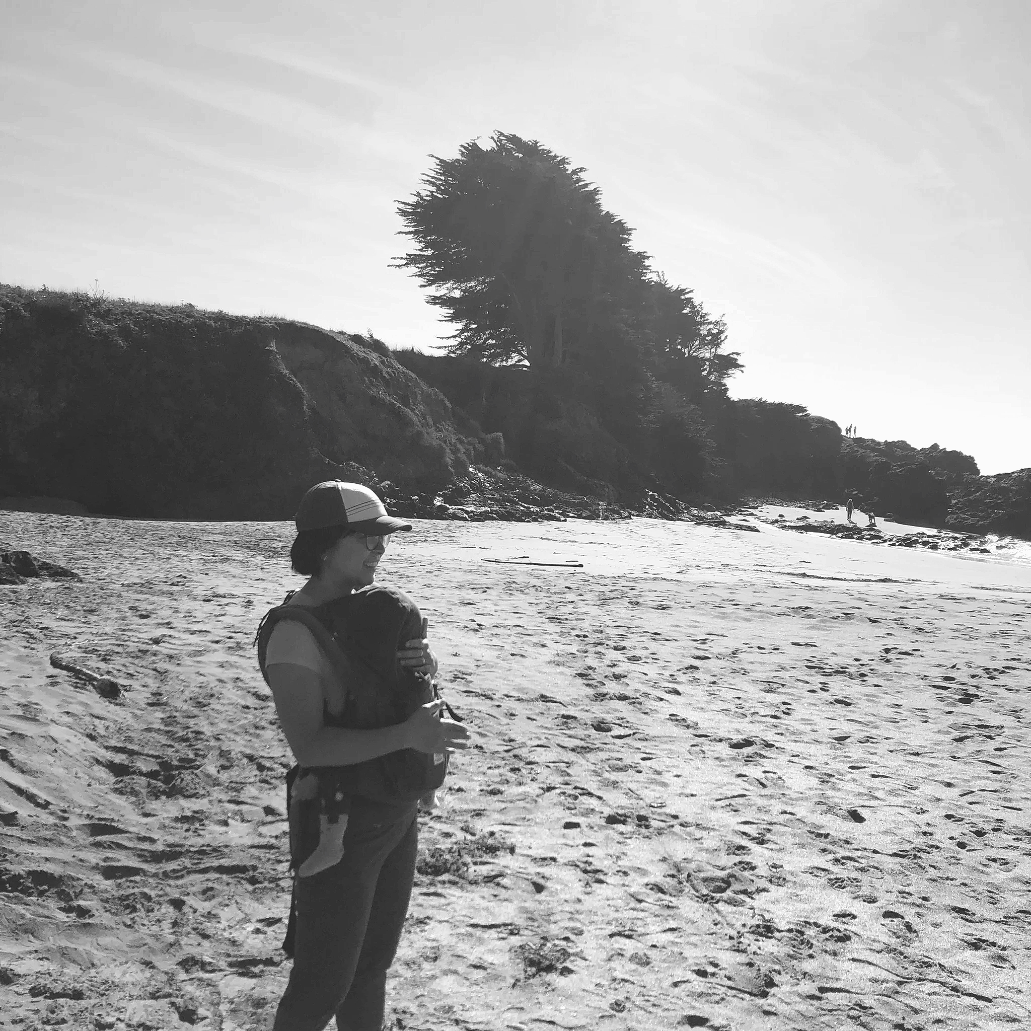 A black and white photo of a beach scene with a woman sitting on the sand, trees on a hill, and a person walking near the water's edge.
