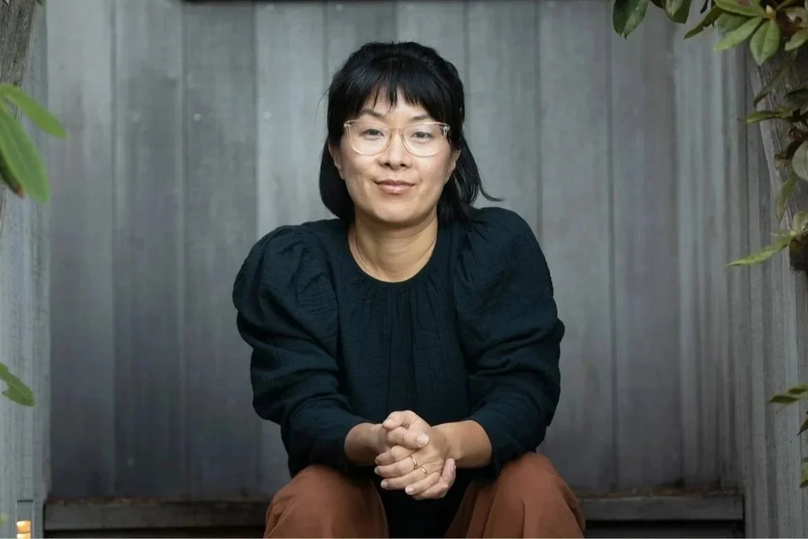 A woman with glasses, short black hair, wearing a black top and brown pants, sits outdoors in front of a wooden fence, with green plants on either side.