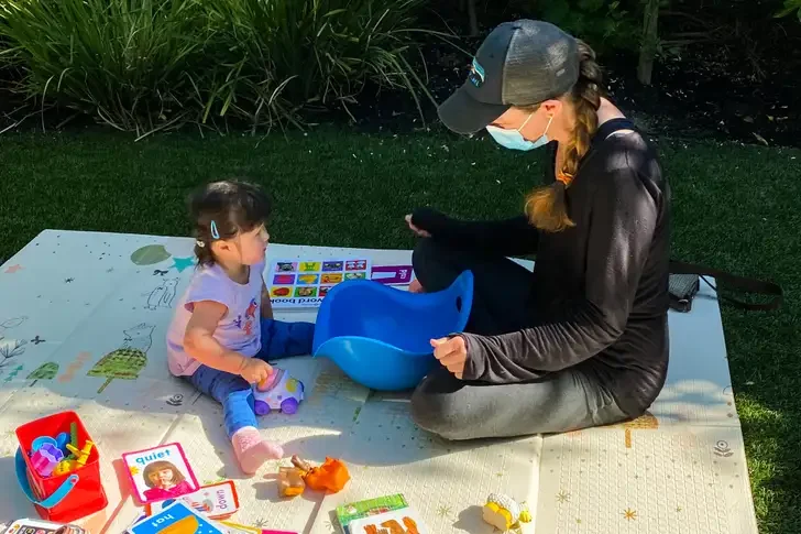 A woman in a face mask and baseball cap sitting on a blanket outdoors, playing with a young girl using toys and educational materials.