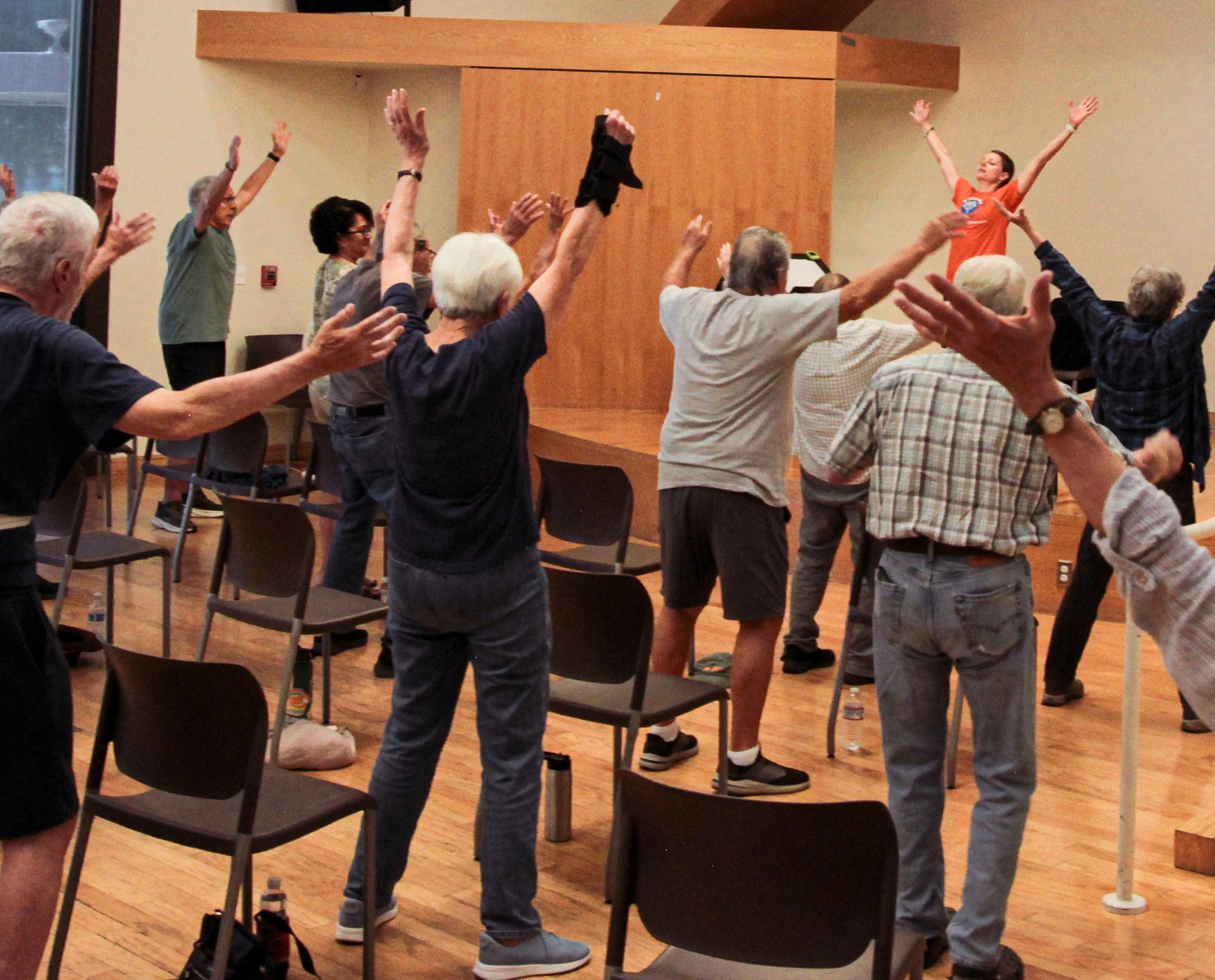 Older adults participating in a seated exercise class, raising their arms and following instructor, in a room with wooden floors and chairs.