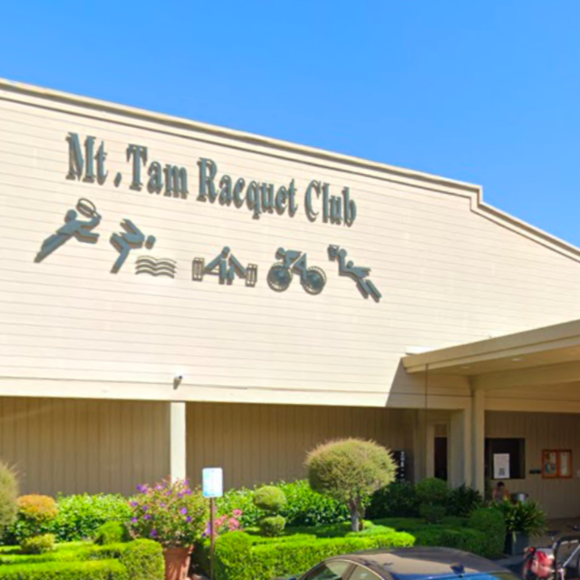 Exterior of the Mt. Tam Racquet Club building with shrubs and a parked car in front, against a clear blue sky.