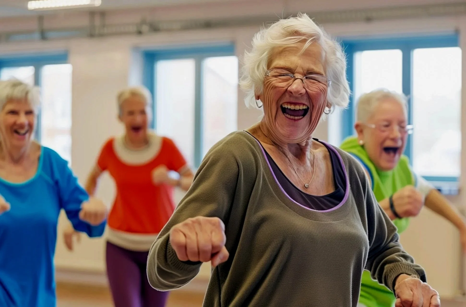 A group of adult women laughing and exercising together in a fitness or community center, with bright windows in the background.