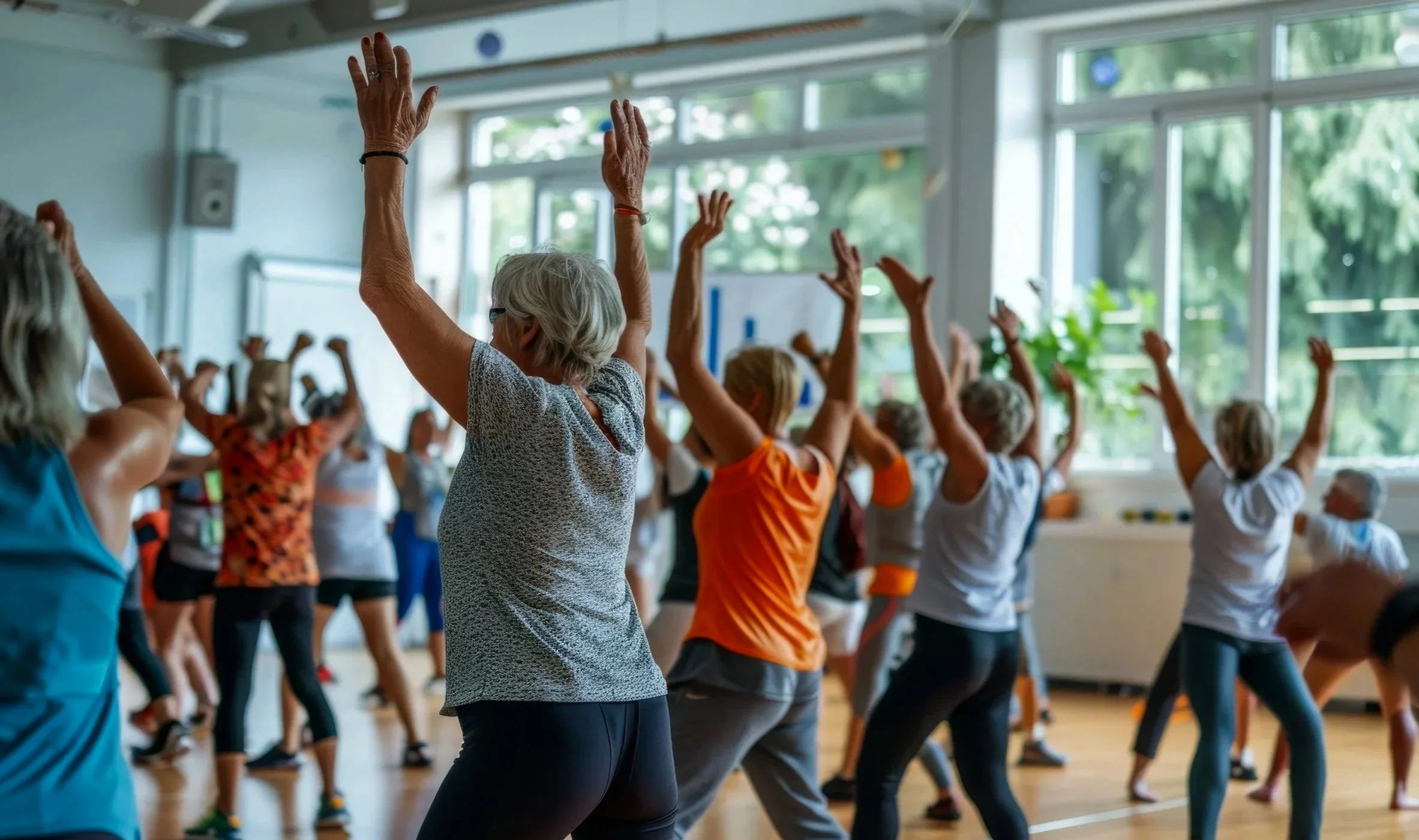 Older adults participating in a dance or fitness class in a brightly lit gym with large windows, some with their arms raised.