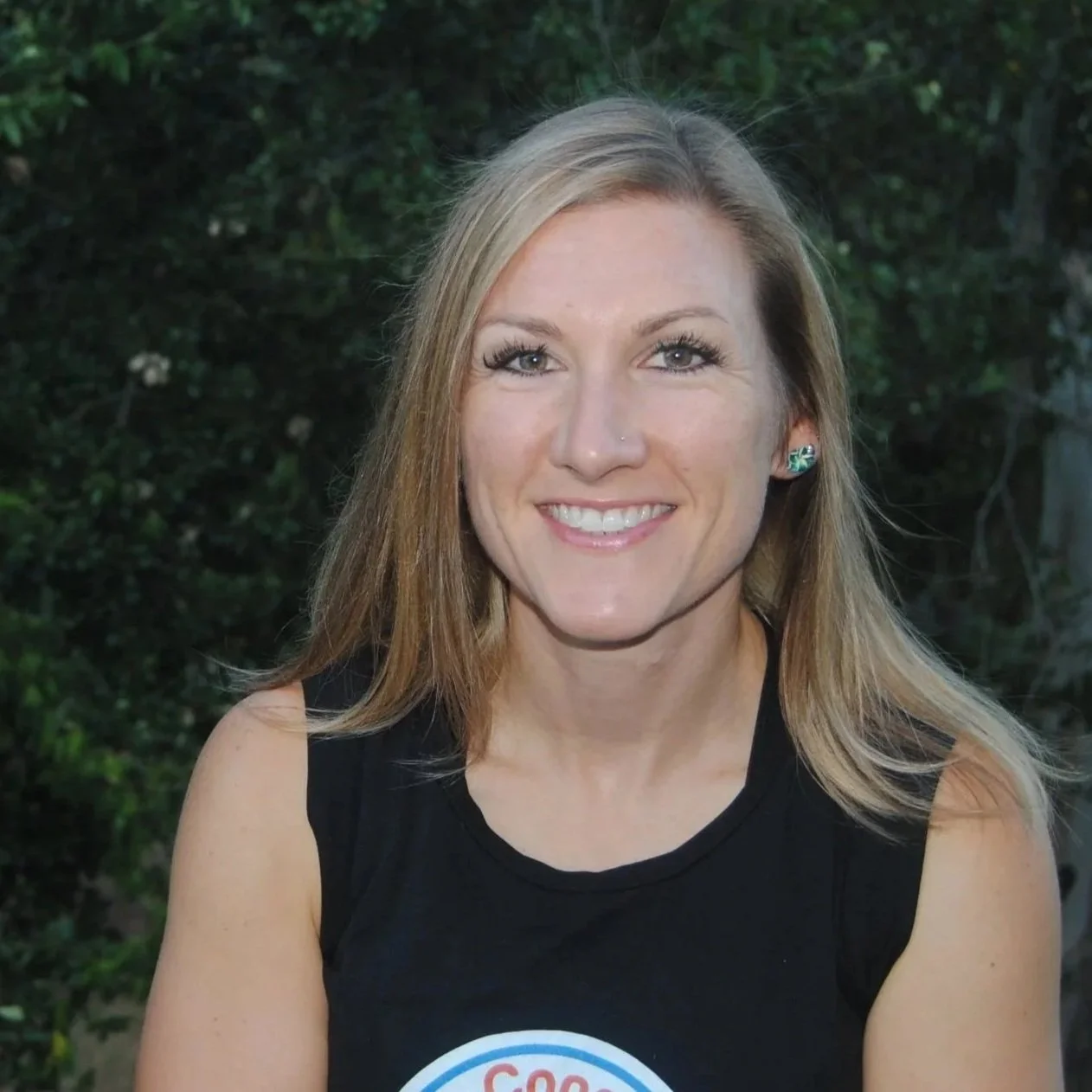 A woman with long blonde hair, wearing a black sleeveless top and turquoise earrings, smiling outdoors with greenery in the background.