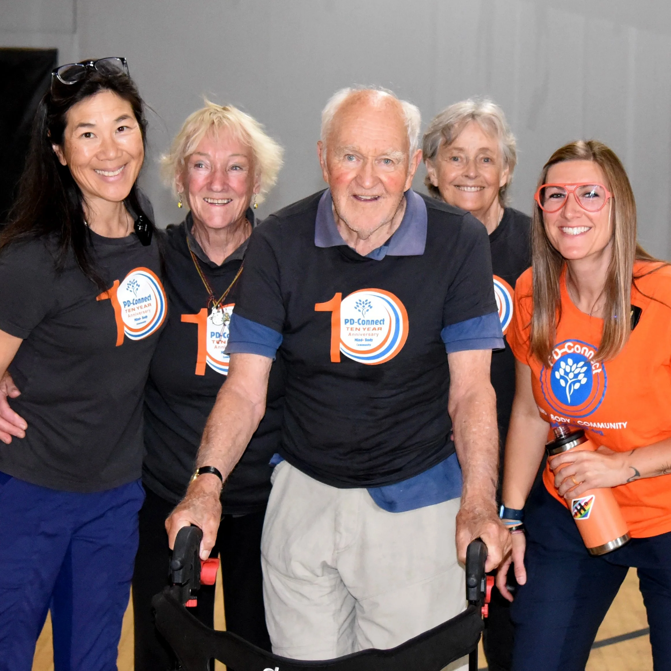 Group of five smiling people at an event, some wearing shirts with a logo celebrating the ten-year anniversary of PD-Connect, standing together with their arms around each other.