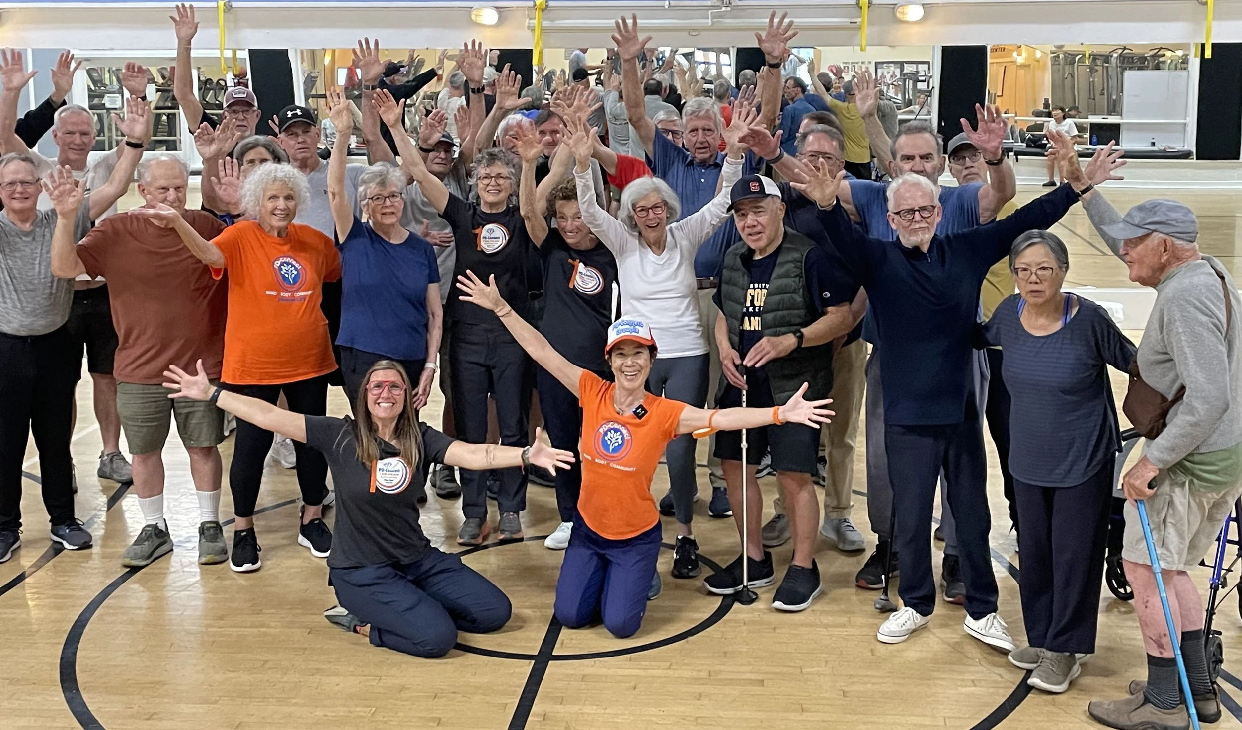 A diverse group of elderly and middle-aged people gathered in a gymnasium, raising their hands and smiling, some wearing sportswear and event badges, with a few in wheelchairs, celebrating together.