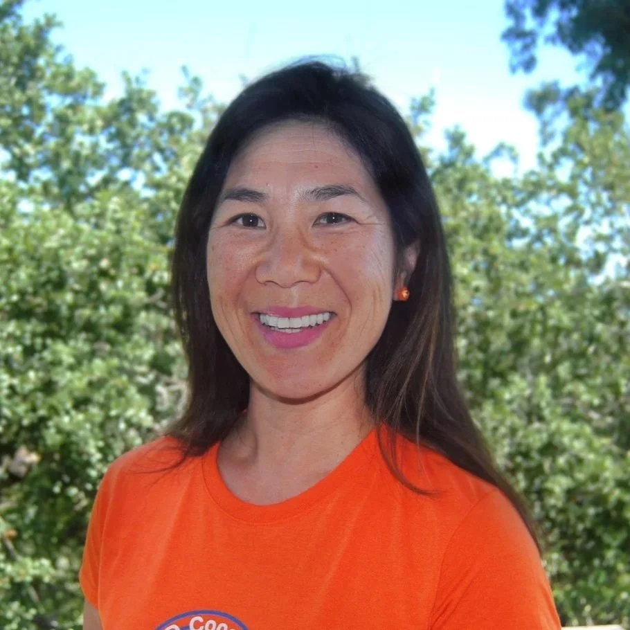 A woman smiling outdoors in front of trees, wearing an orange T-shirt and orange earrings.