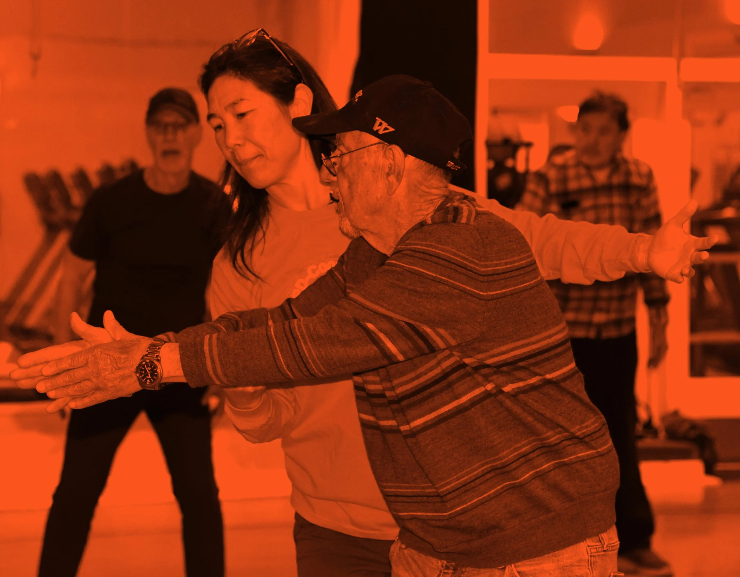 A man and an instructor together, with the woman slightly holding the man's arm, in a lively class with other people in the background.