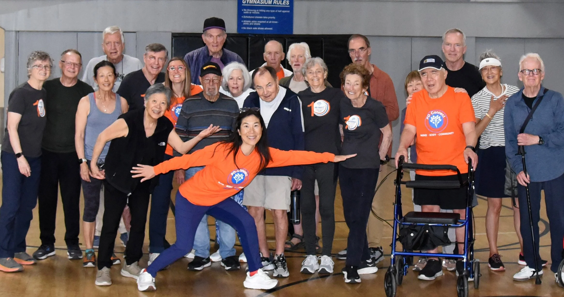 Group of elderly and middle-aged people gathered in a gymnasium, some wearing sportswear and a few with walkers, celebrating an event related to Parkinson's disease awareness, as indicated by the shirts and badges. A woman in an orange shirt is strik