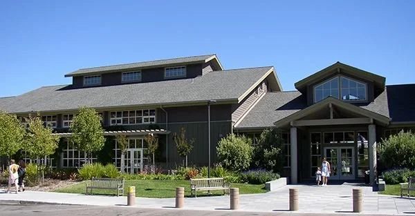A large, modern building with gray siding and numerous windows, surrounded by greenery and trees, with people walking near the entrance on a sunny day.