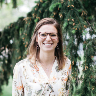 A woman with glasses and earrings smiling outdoors in front of a large green tree.