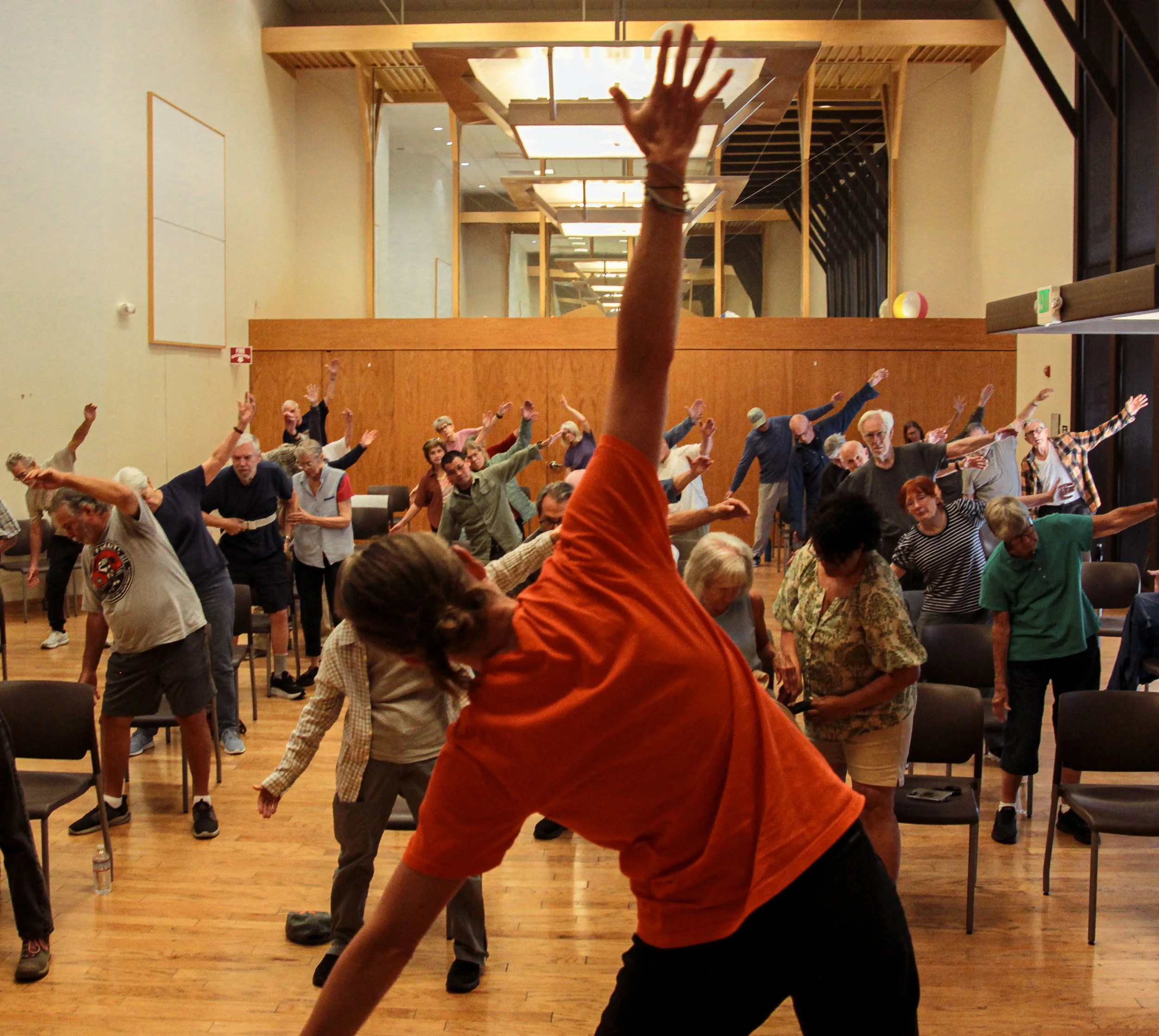 A large group of people participating in a dance or exercise class in a room with wooden floors and modern ceiling lights, some with their arms raised and others bent or extended, with a raised arm visible in the foreground.