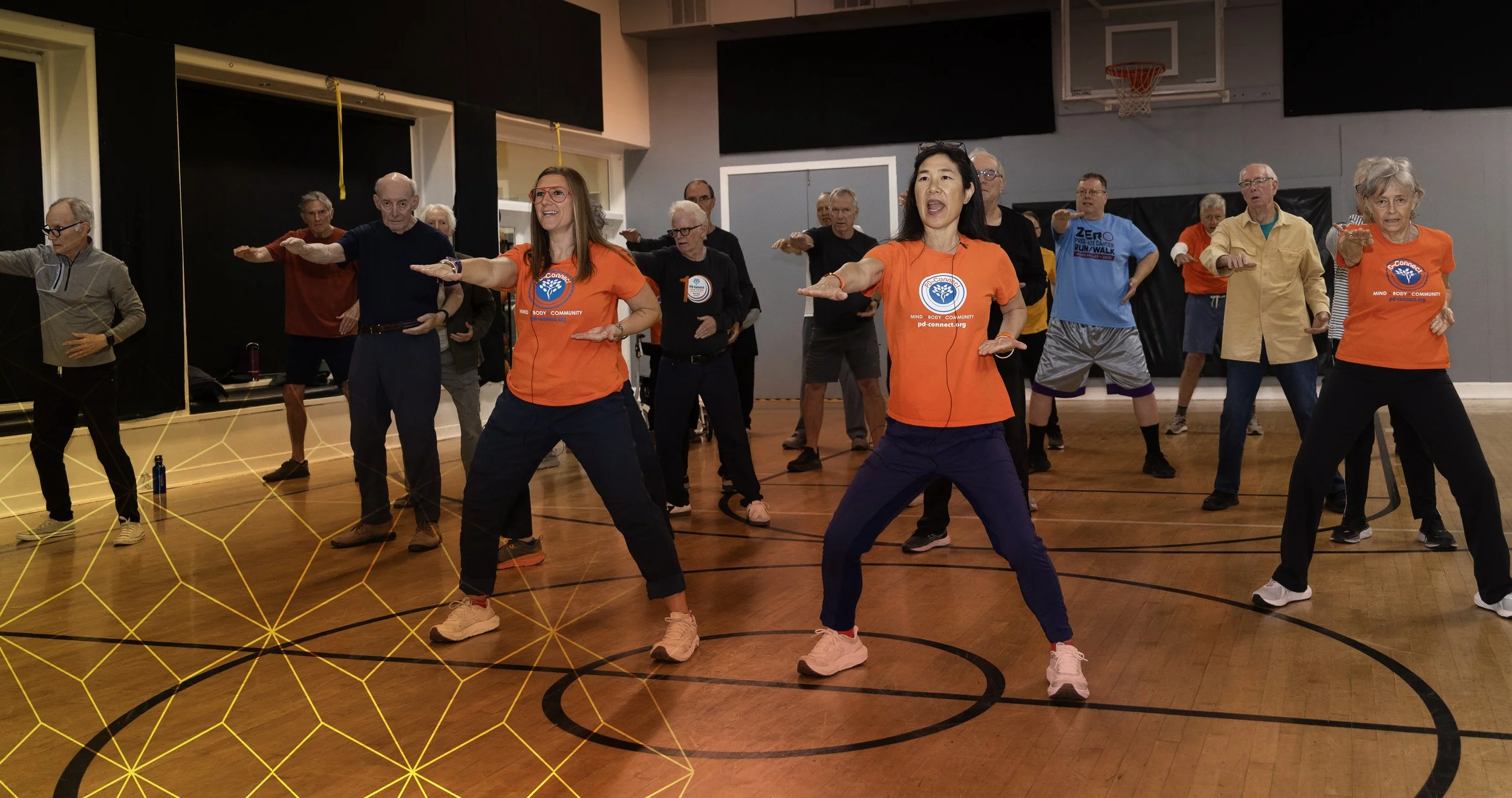 A group of elderly people participating in a fitness class, led by an instructor, inside a gymnasium. They are performing a stretching exercise with their arms extended forward.
