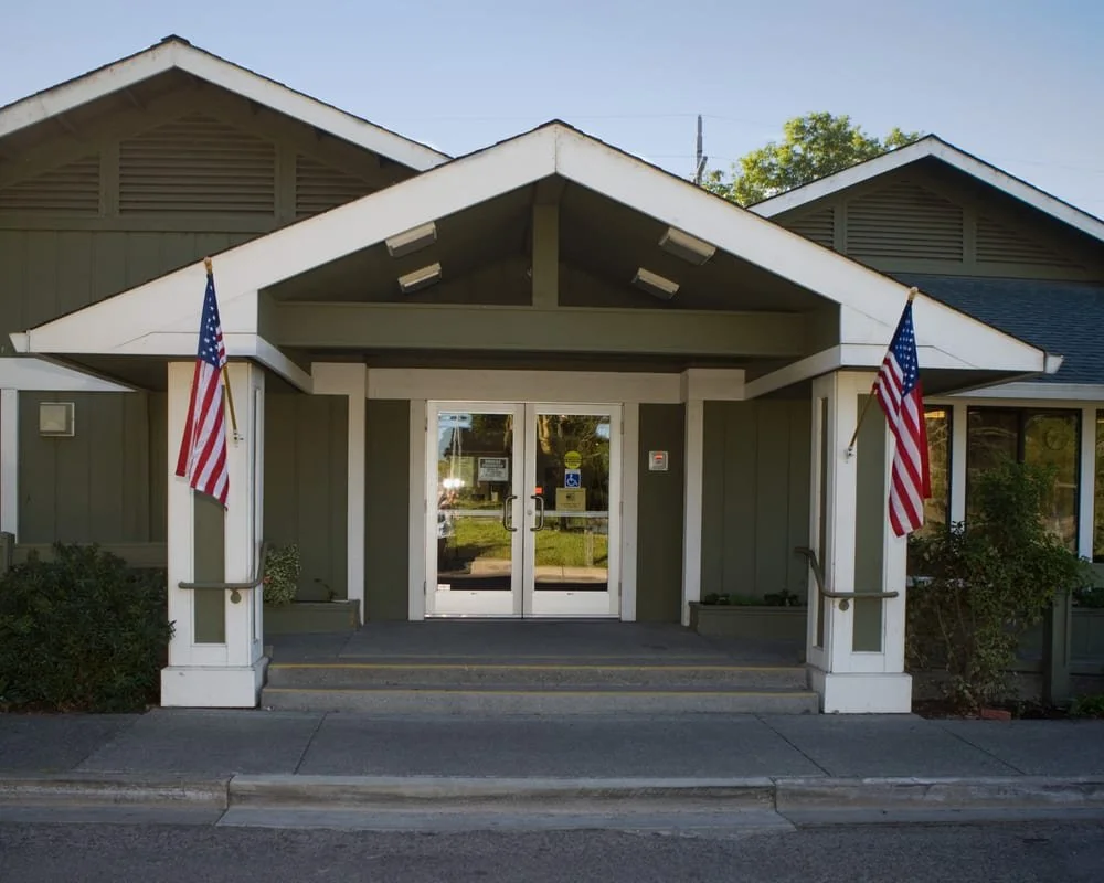 Entrance of a building with American flags on each side, glass double doors, small steps, and a covered porch.