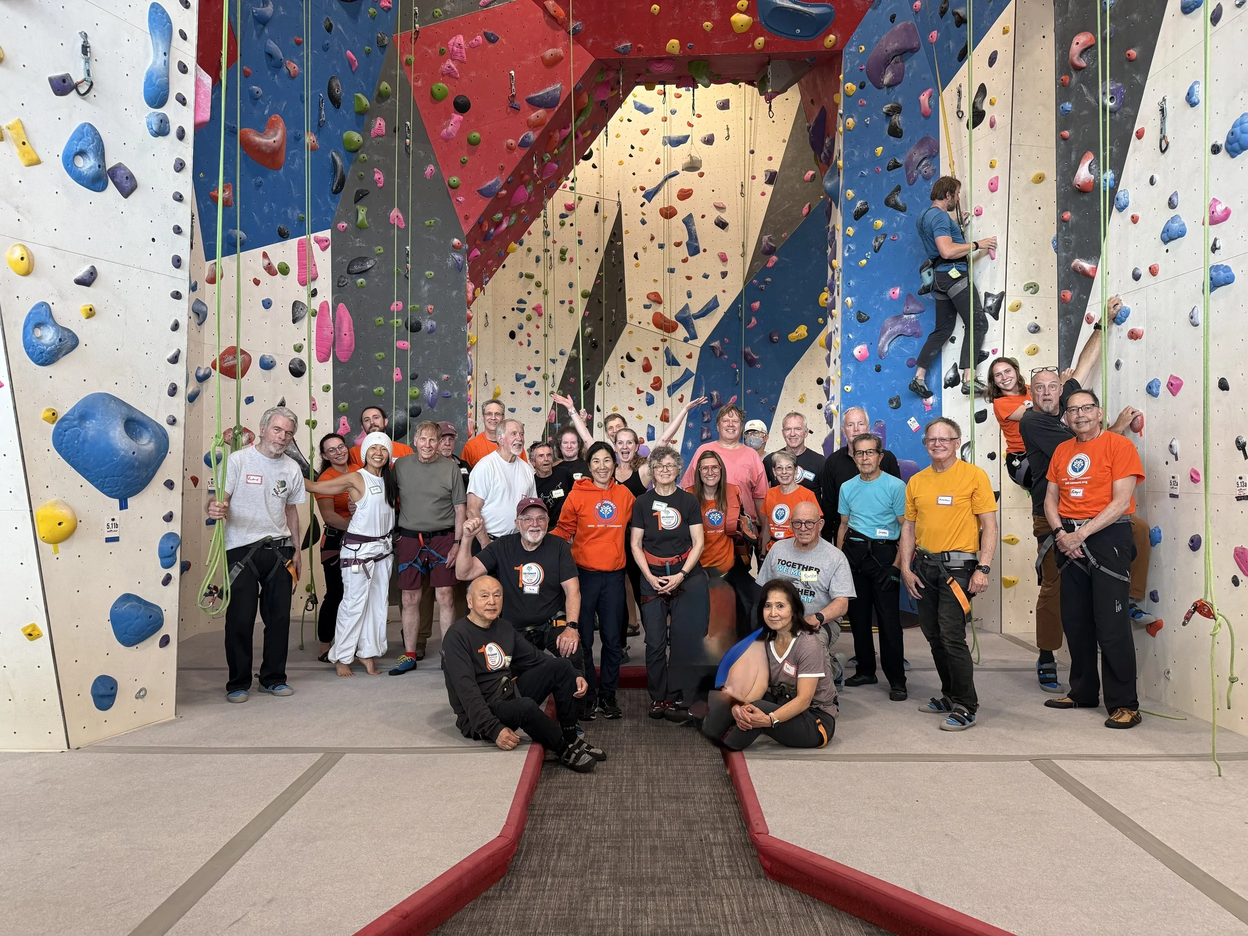 A group of people posing in front of an indoor rock climbing wall with various colored holds. The team appears happy and are wearing casual sportwear, some with climbing harnesses, with a few individuals making celebratory gestures.