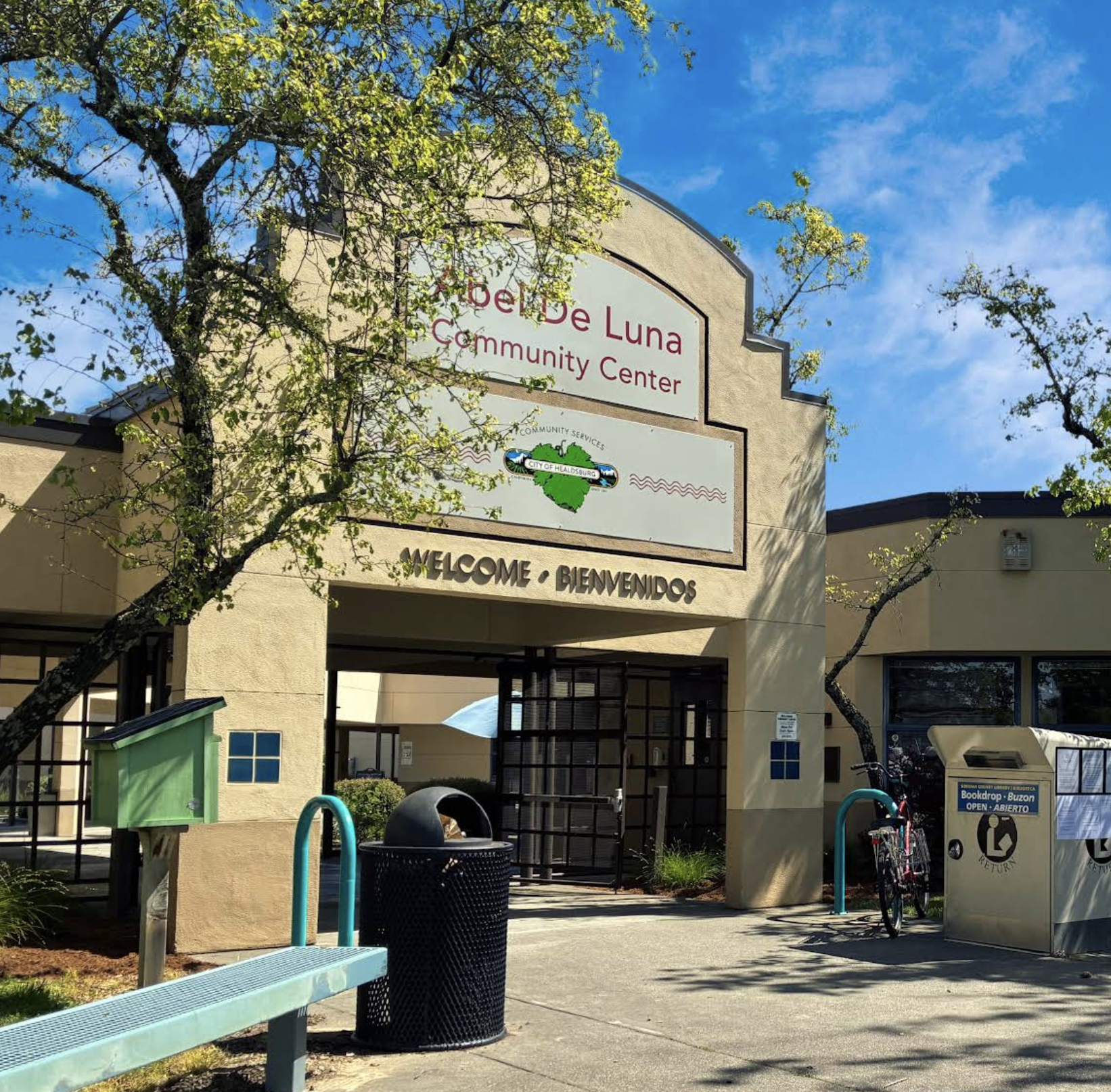 The exterior of the Abel De Luna Community Center, featuring beige walls, a sign with the center's name, and a blue sky with some clouds. There is a bench, trash can, bike rack with bicycles, and a book drop box outside. Green trees and some shadows are also visible.