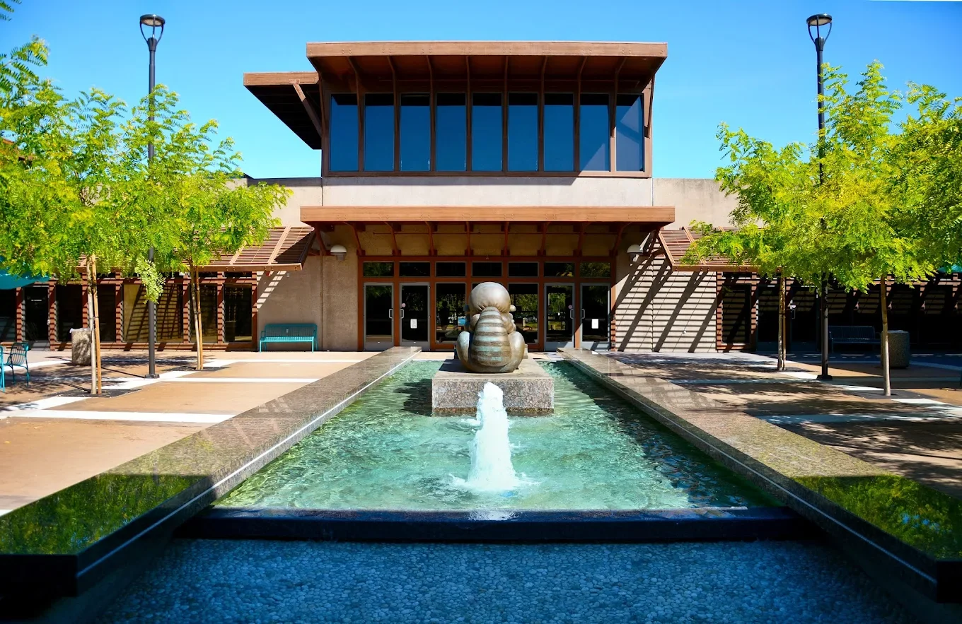 Front view of a modern building with a water fountain in the foreground, surrounded by trees and benches.