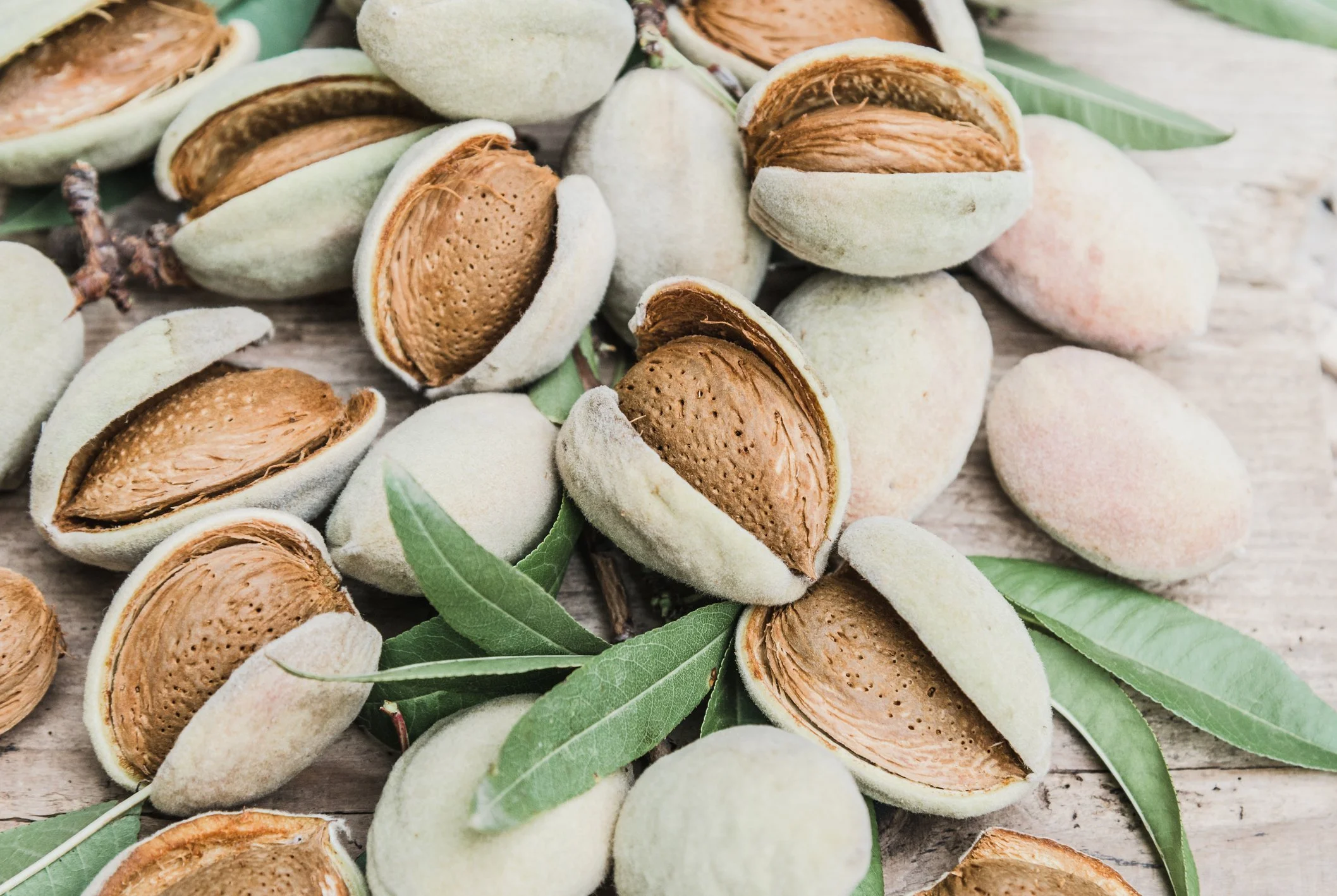 Close-up of several open and closed almonds with green leaves scattered around, placed on a wooden surface.