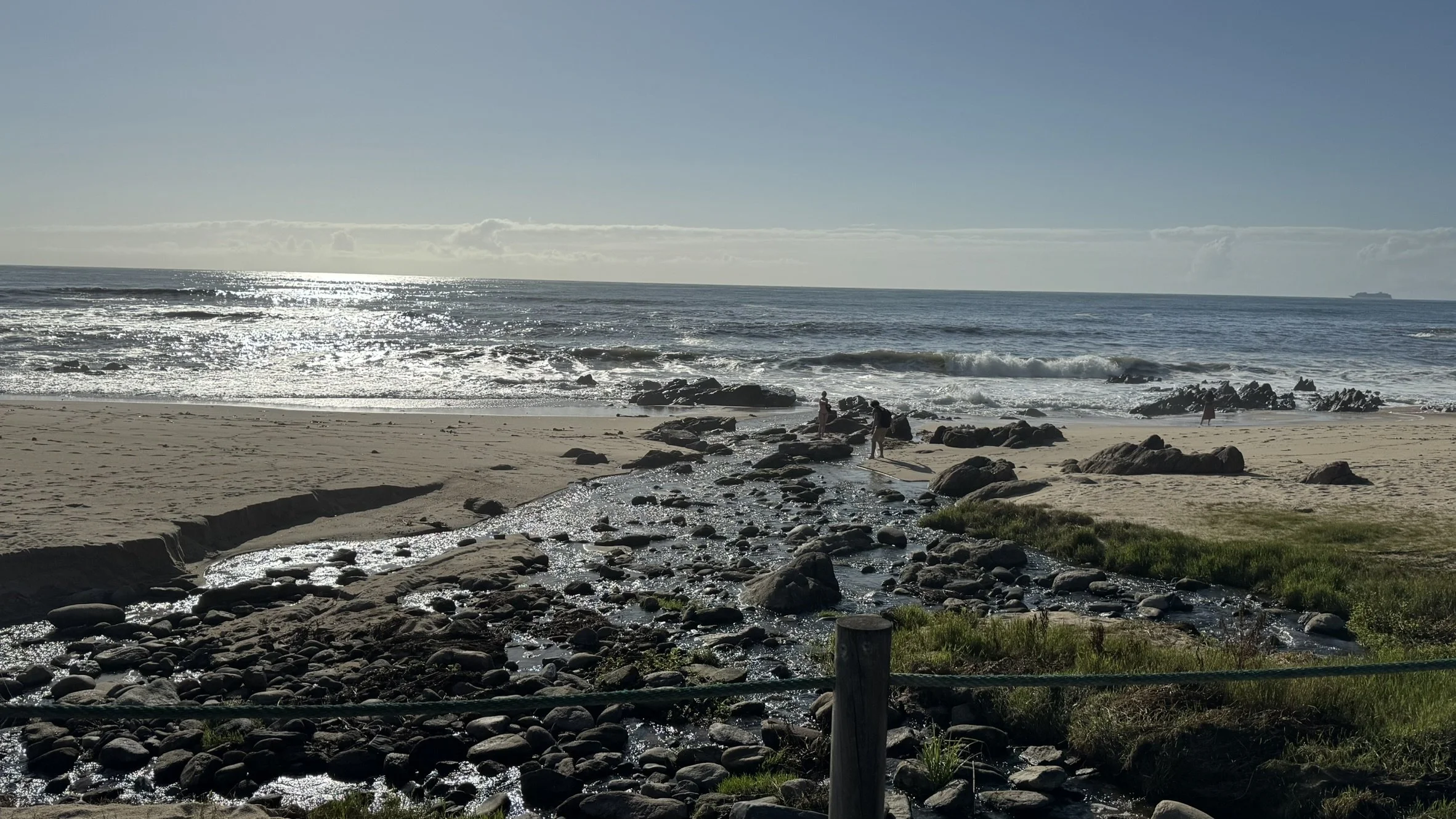 A beach with rocks, a small stream flowing into the ocean, and a few people near the rocks, with the sun reflecting off the water and a ship on the horizon.