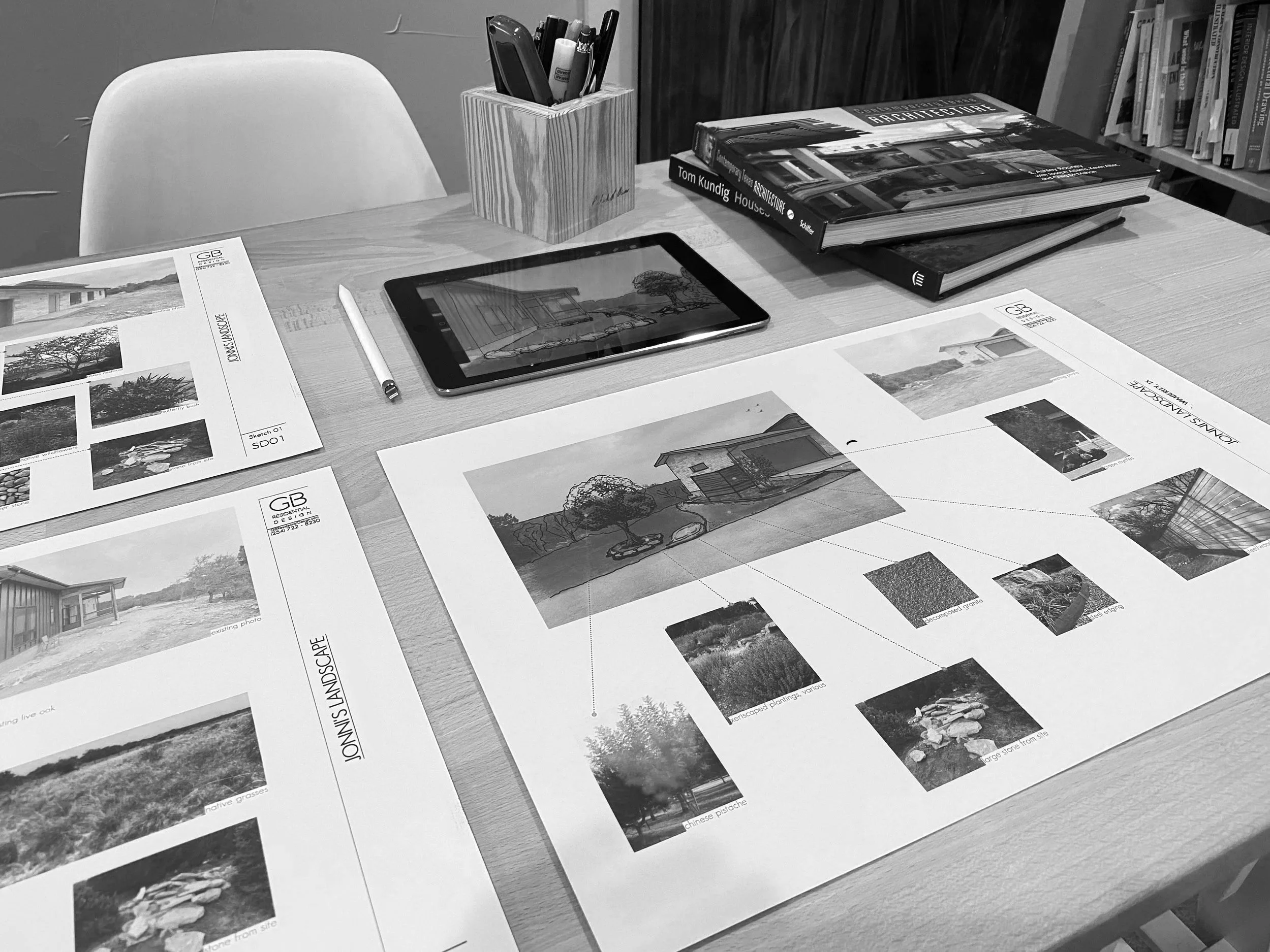 A desk with architectural and landscape design plans, a tablet displaying a house sketch, three large architecture books, a white pen, and a wooden pen holder with various pens and markers.