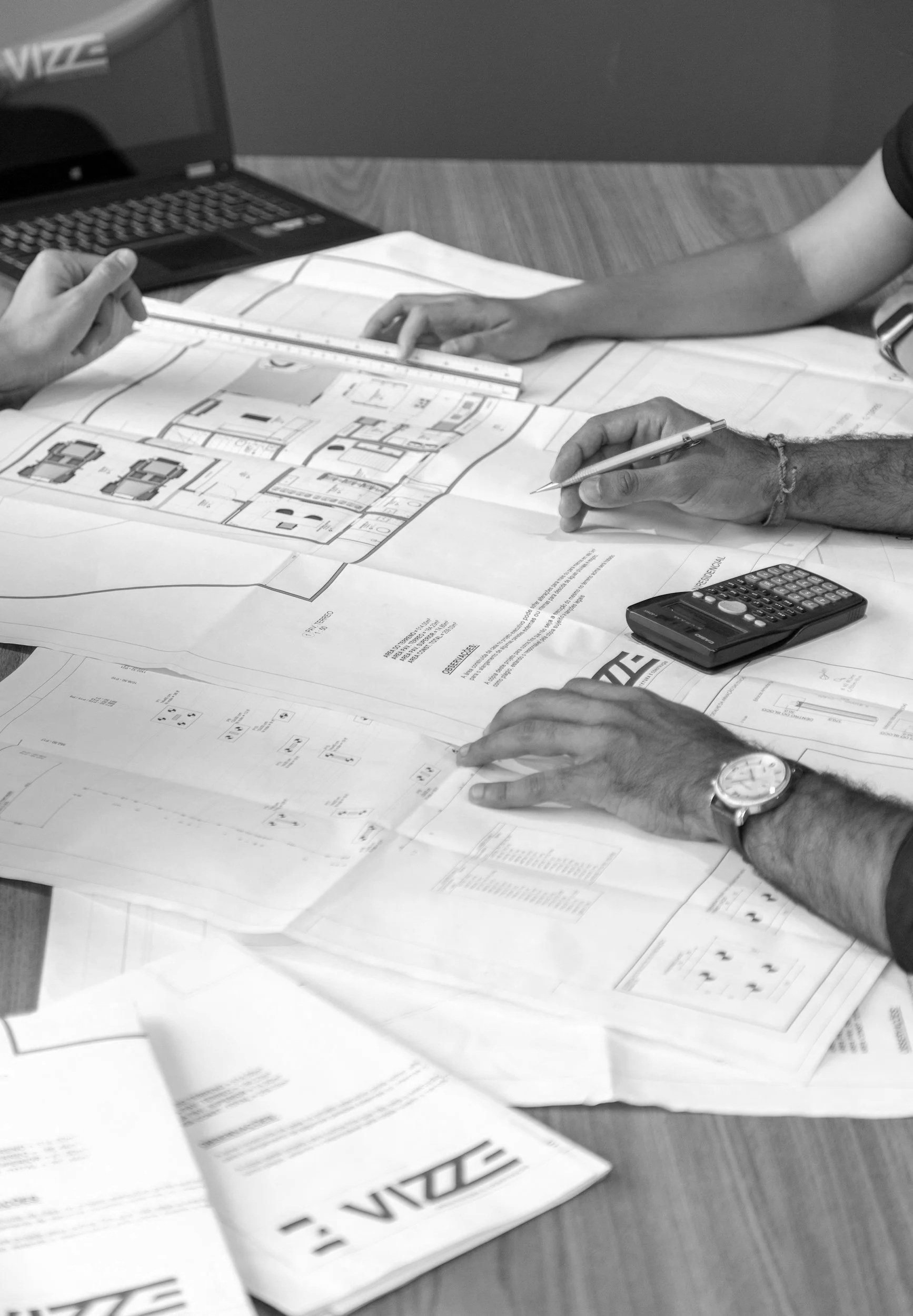 Two people working on architectural blueprints spread out on a table; one person is writing, the other is holding a scale ruler; a laptop and calculator are also on the table.