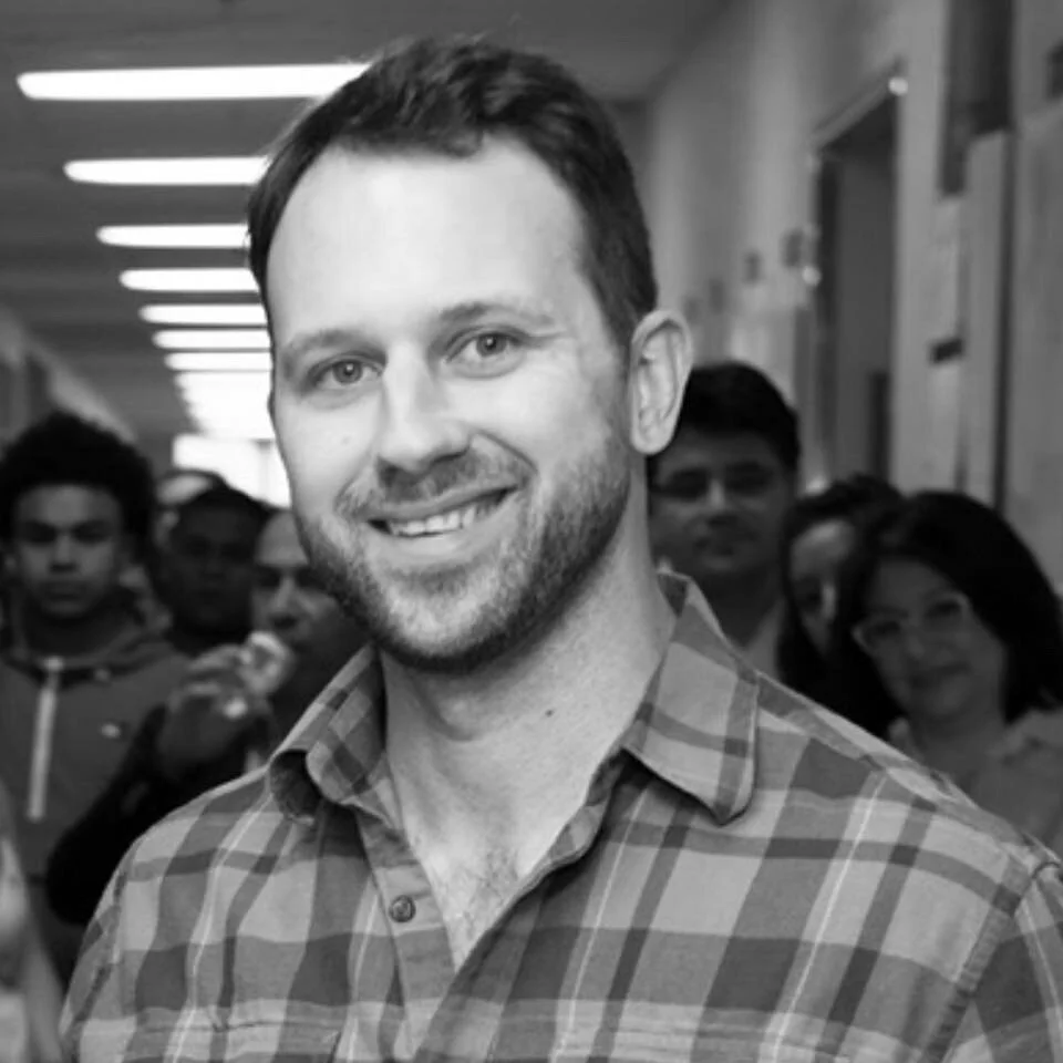 A smiling man with a beard and short hair in a checkered shirt, standing in a hallway with multiple people in the background.