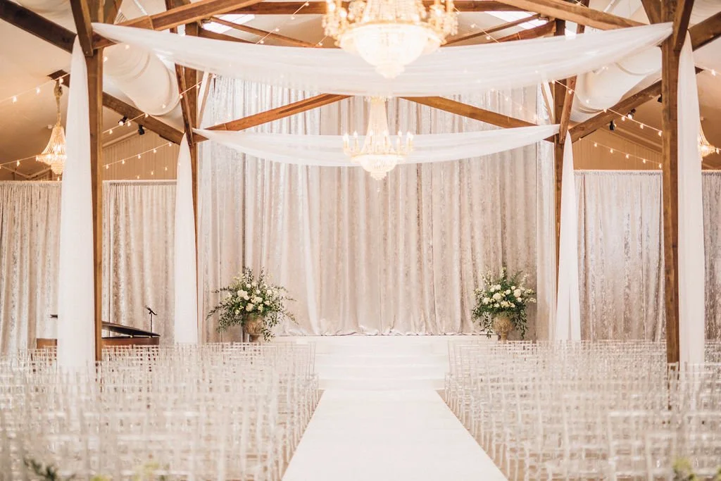 Elegant wedding ceremony setup with wooden beams, draped white fabric, floral arrangements, and chandelier lighting in Pensacola, Florida.