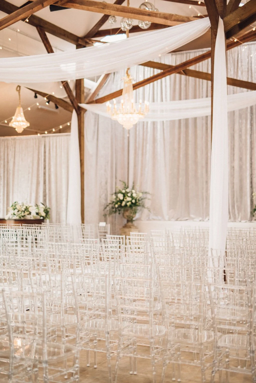 Elegant wedding ceremony setup with white chairs, draped white fabric, chandeliers, and floral arrangements inside a wooden-beamed venue in Pensacola