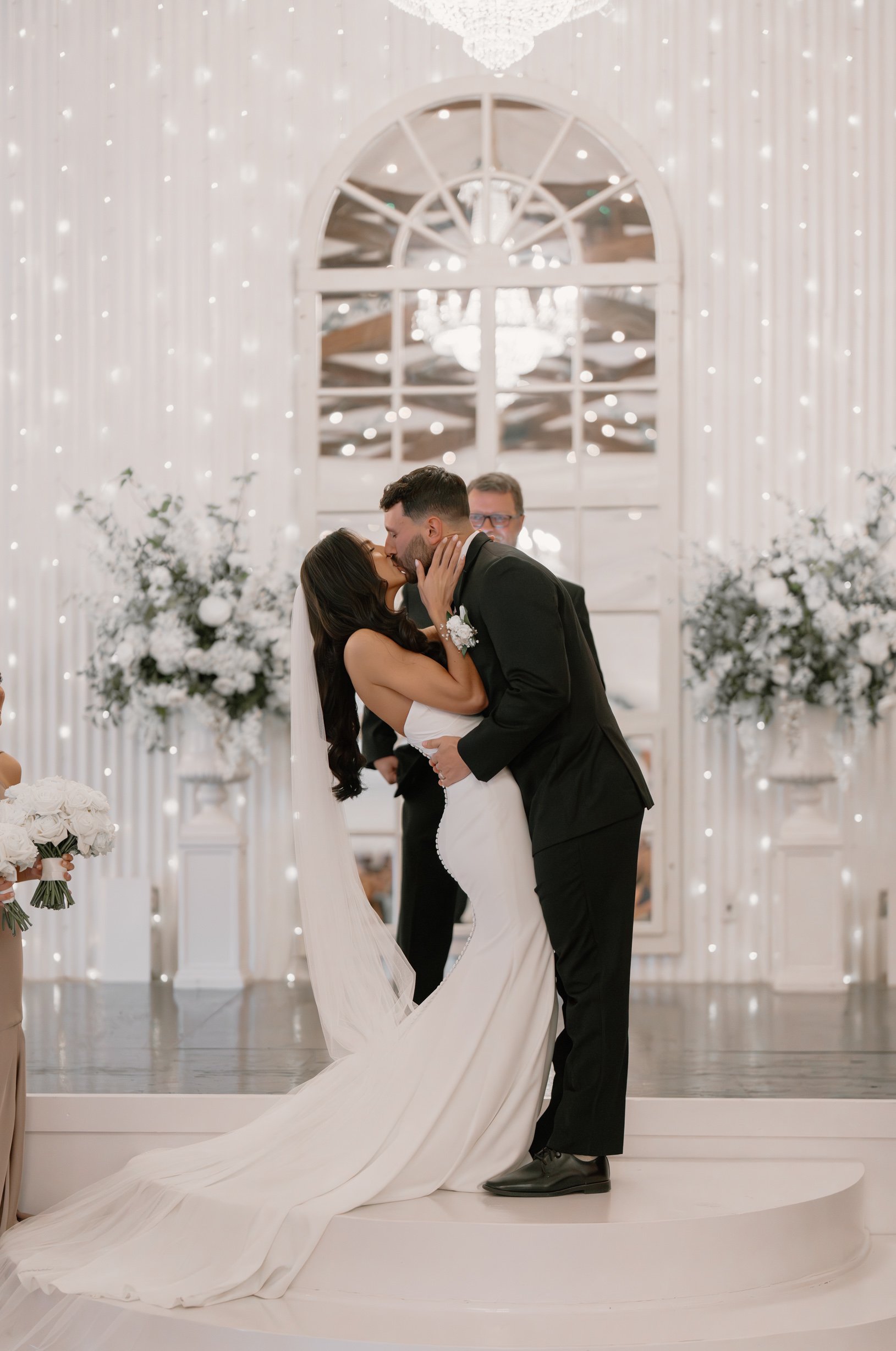 A couple getting married, sharing a kiss during their wedding ceremony in an elegant, white decorated venue with floral arrangements and string lights.