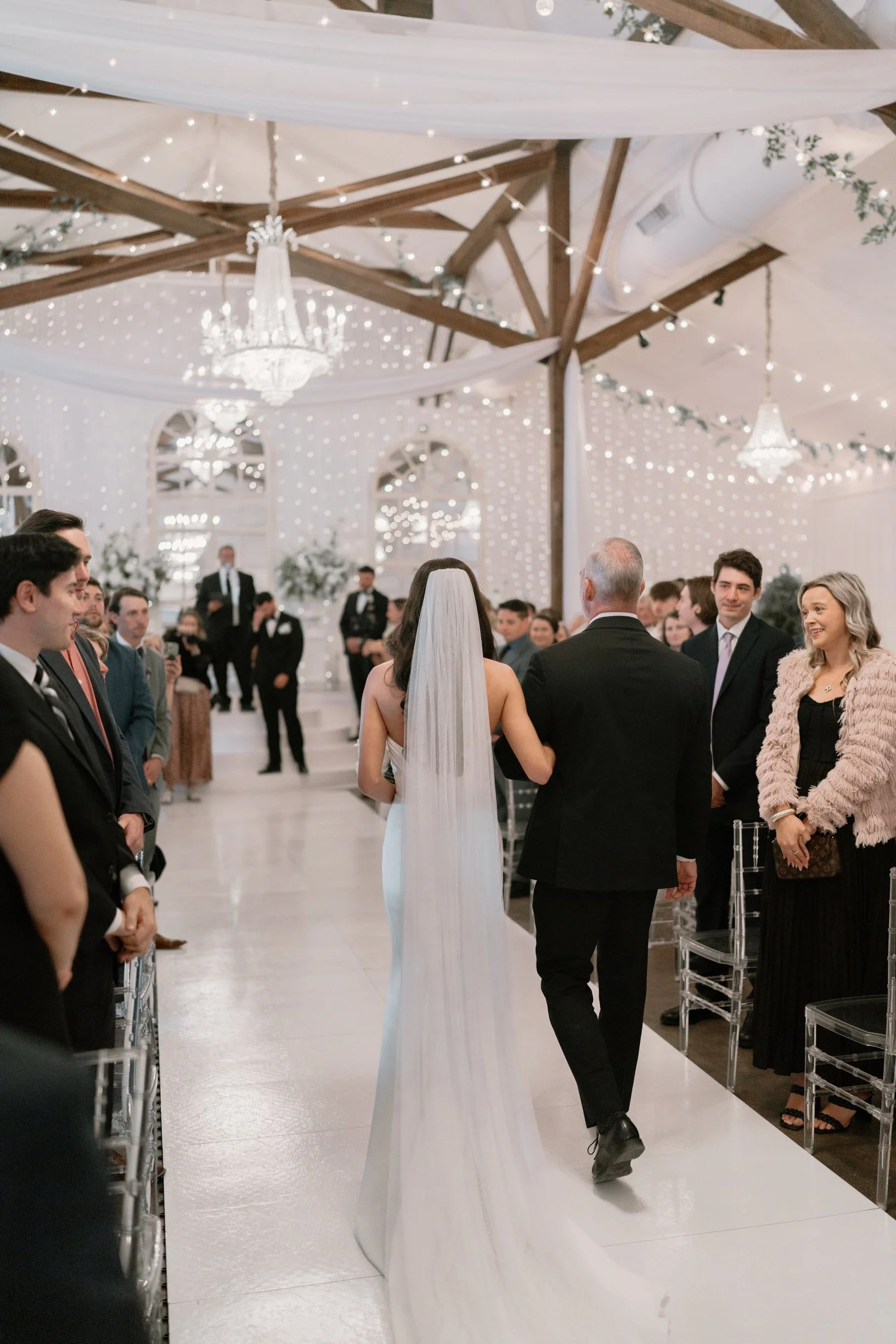 Bride walking down the aisle at her wedding ceremony with her father, as guests and wedding party look on inside a decorated Pensacola wedding venue with chandeliers and string lights.